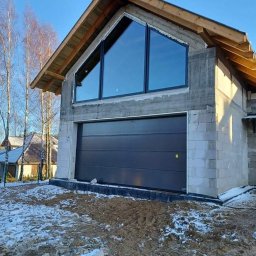 Pentra Plus - Dom w trakcie budowy z zamontowaną ciemnoszarą bramą garażową segmentową i dużym trójkątnym oknem z czarną ramą na szczycie, widoczne bloczki betonowe i ośnieżona ziemia.