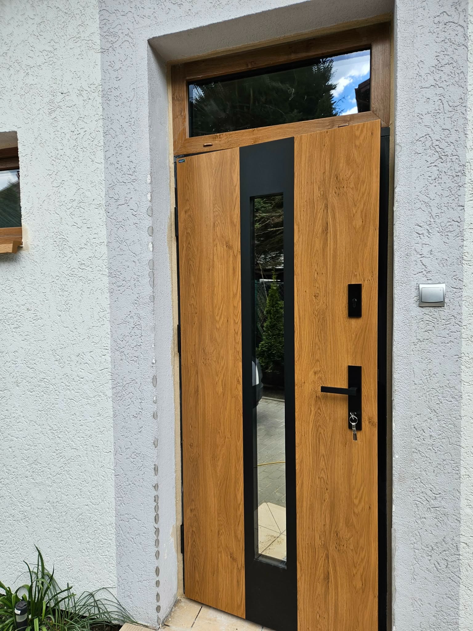 Drewniane drzwi wejściowe z czarnymi elementami i szybą, zamontowane w jasnym budynku z oknem nad drzwiami. Widoczne klucze w zamku i fragment roślinności u dołu.