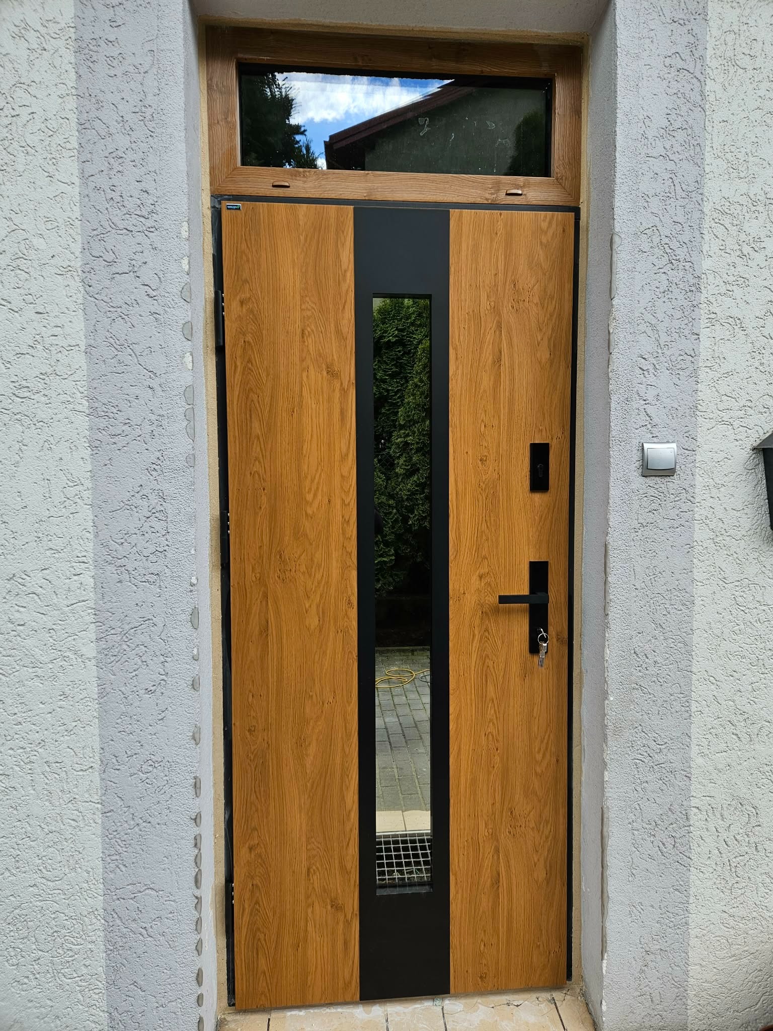 Zewnętrzne drzwi wejściowe z drewnopodobnym panelem i czarnymi elementami, osadzone w szarej elewacji z widocznym oknem nad drzwiami i kluczami w zamku.