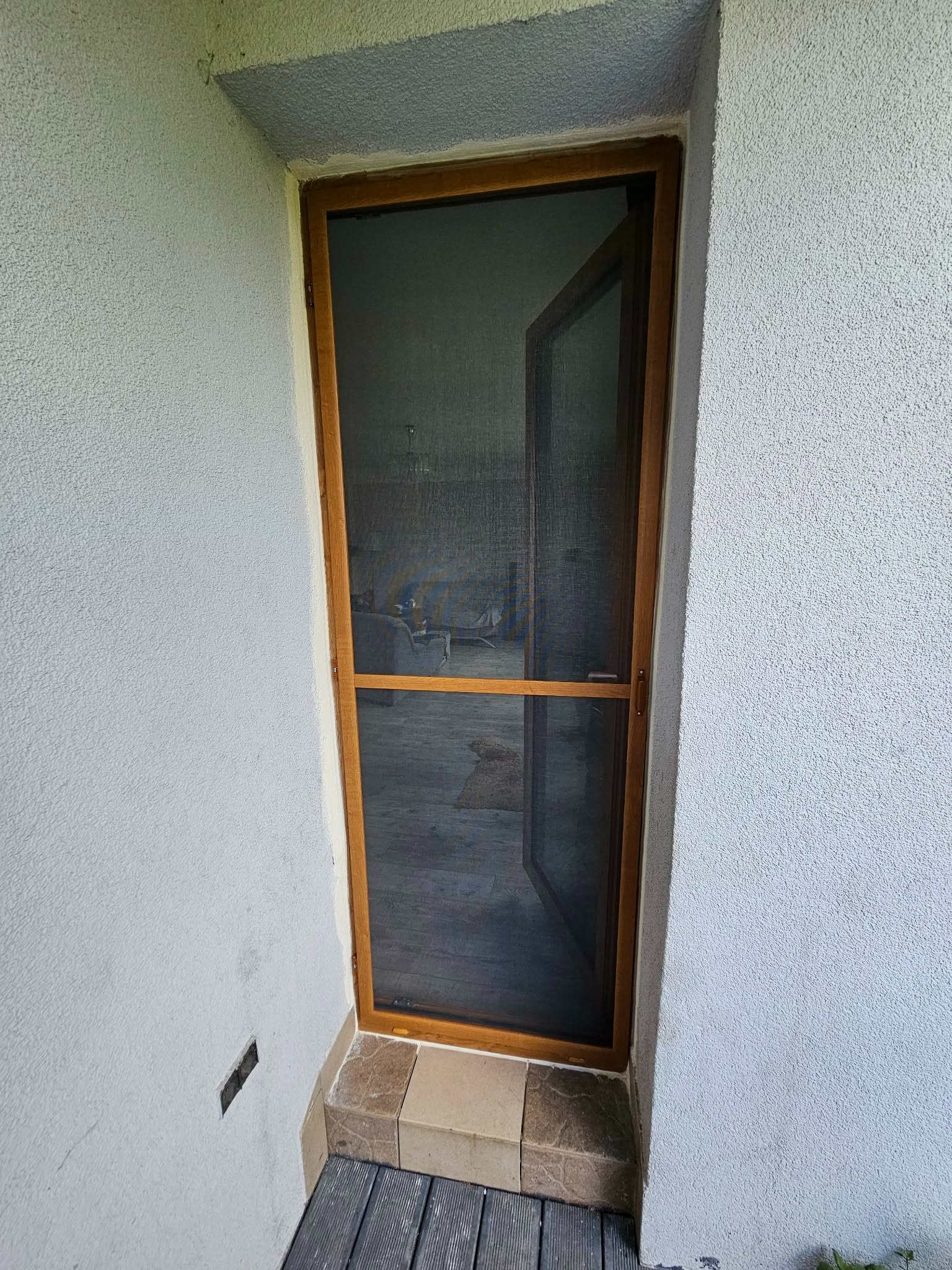 Otwarta moskitiera w drewnianej ramie, zamontowana w wejściu do budynku z szarą, teksturowaną elewacją. Widoczne płytki na progu i fragment drewnianego podestu.