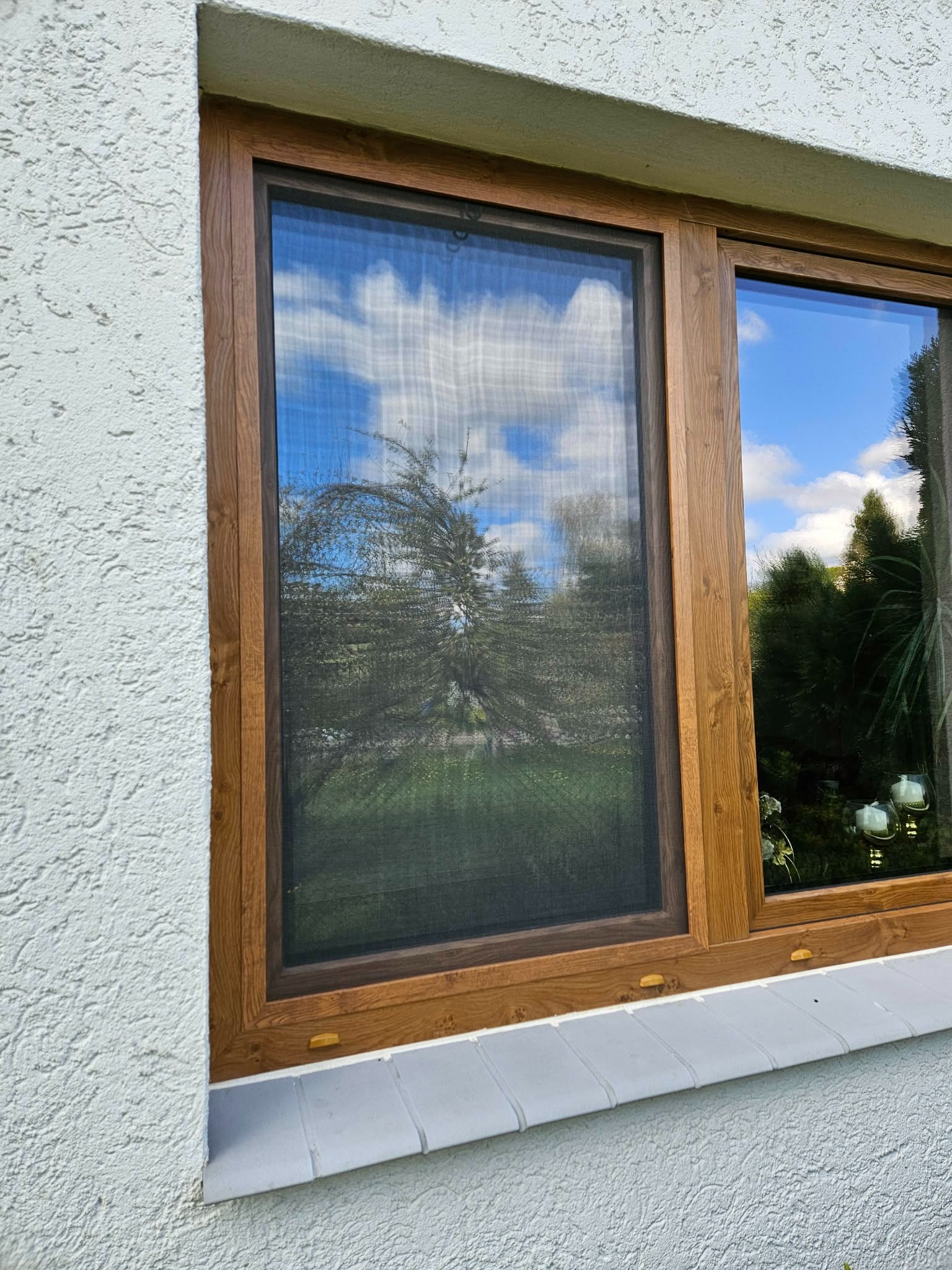 Drewnopodobne ramy okienne z moskitierą, odbijające błękitne niebo i zieleń ogrodu. Biała, teksturowana ściana zewnętrzna i szary parapet dodają kontrastu.