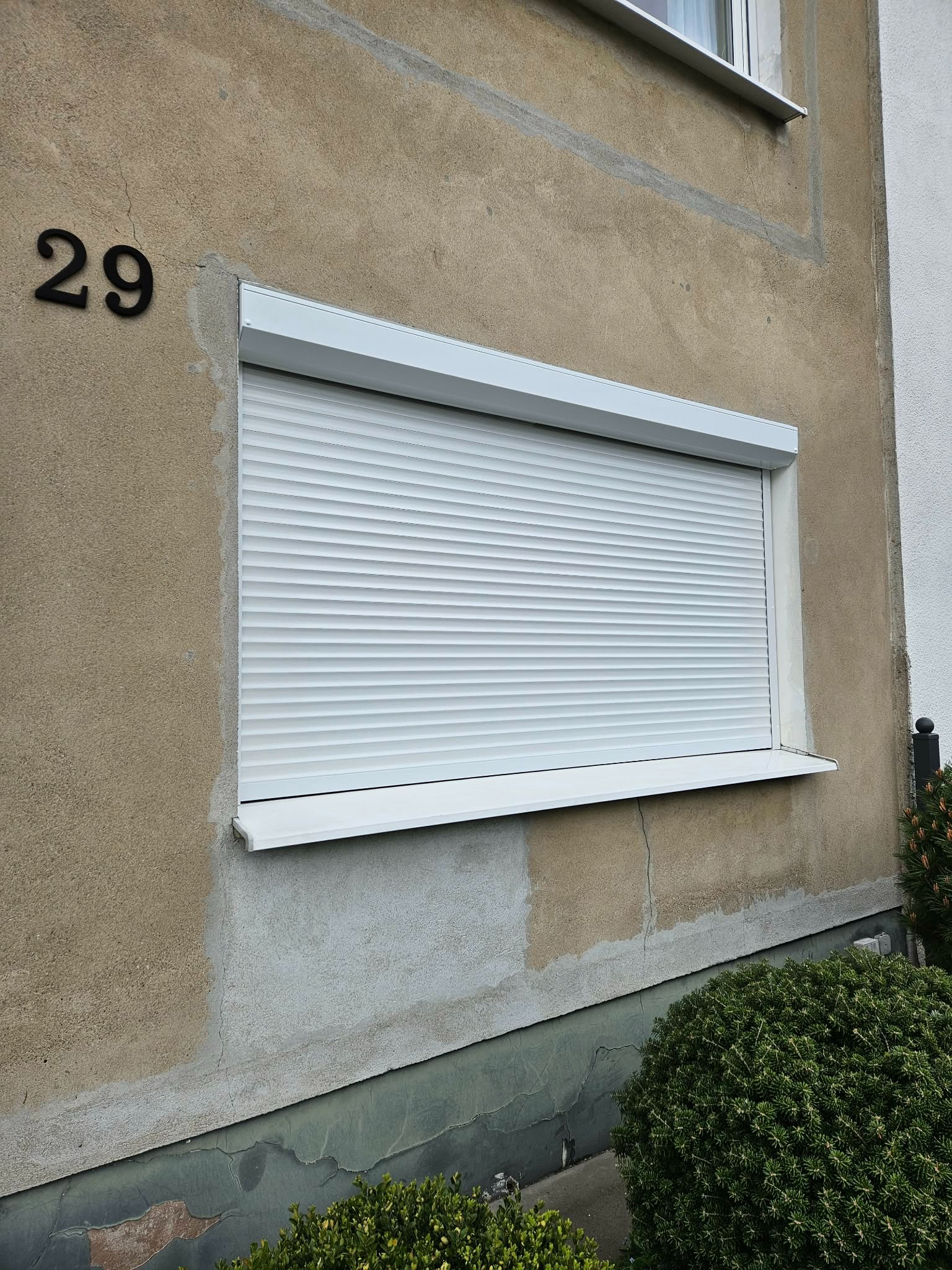 Biała roleta zewnętrzna na oknie budynku z numerem 29, widoczna z poziomu gruntu. Roleta opuszczona, zasłania okno. Tynk w odcieniach beżu i szarości.