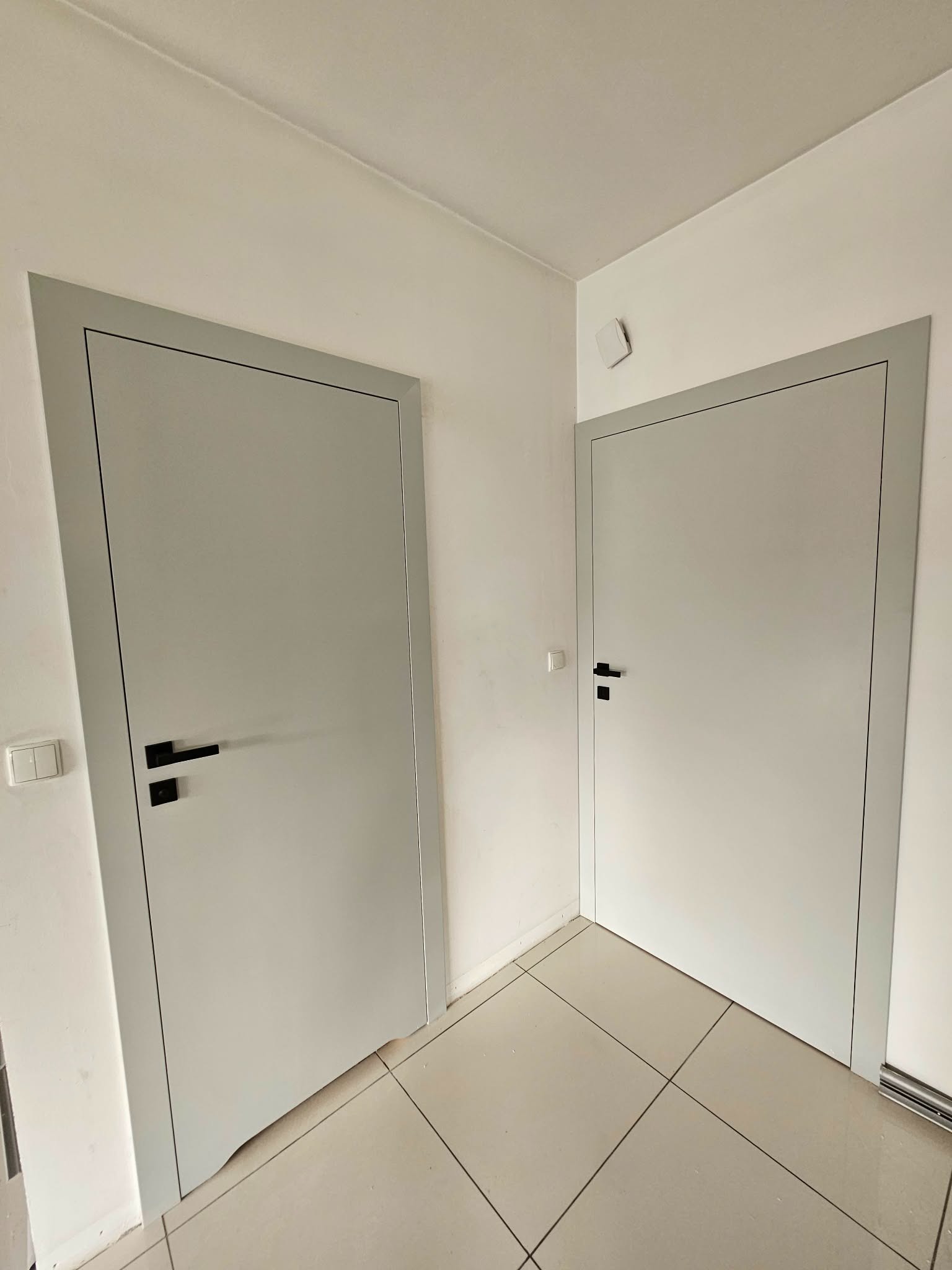 Dwa białe drzwi wewnętrzne z szarą ościeżnicą i czarnymi klamkami w korytarzu z jasnymi płytkami podłogowymi. Minimalistyczny design.