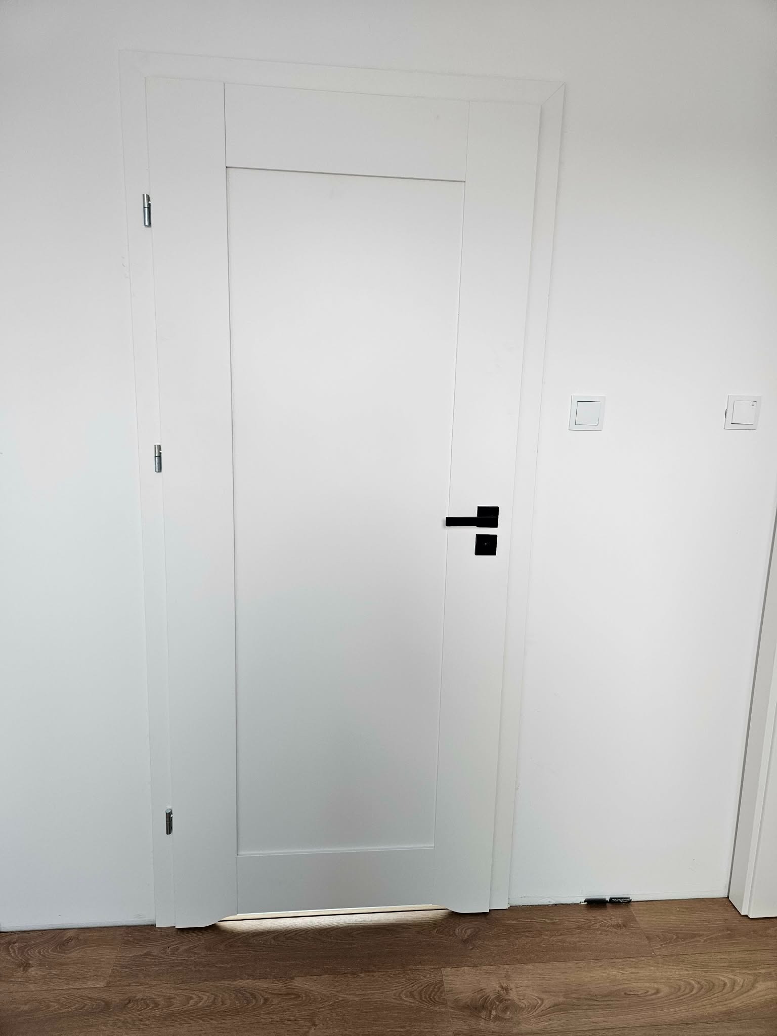 Minimalistyczne, białe drzwi wewnętrzne z czarną klamką na tle białej ściany z włącznikami. Widoczny fragment podłogi z paneli.