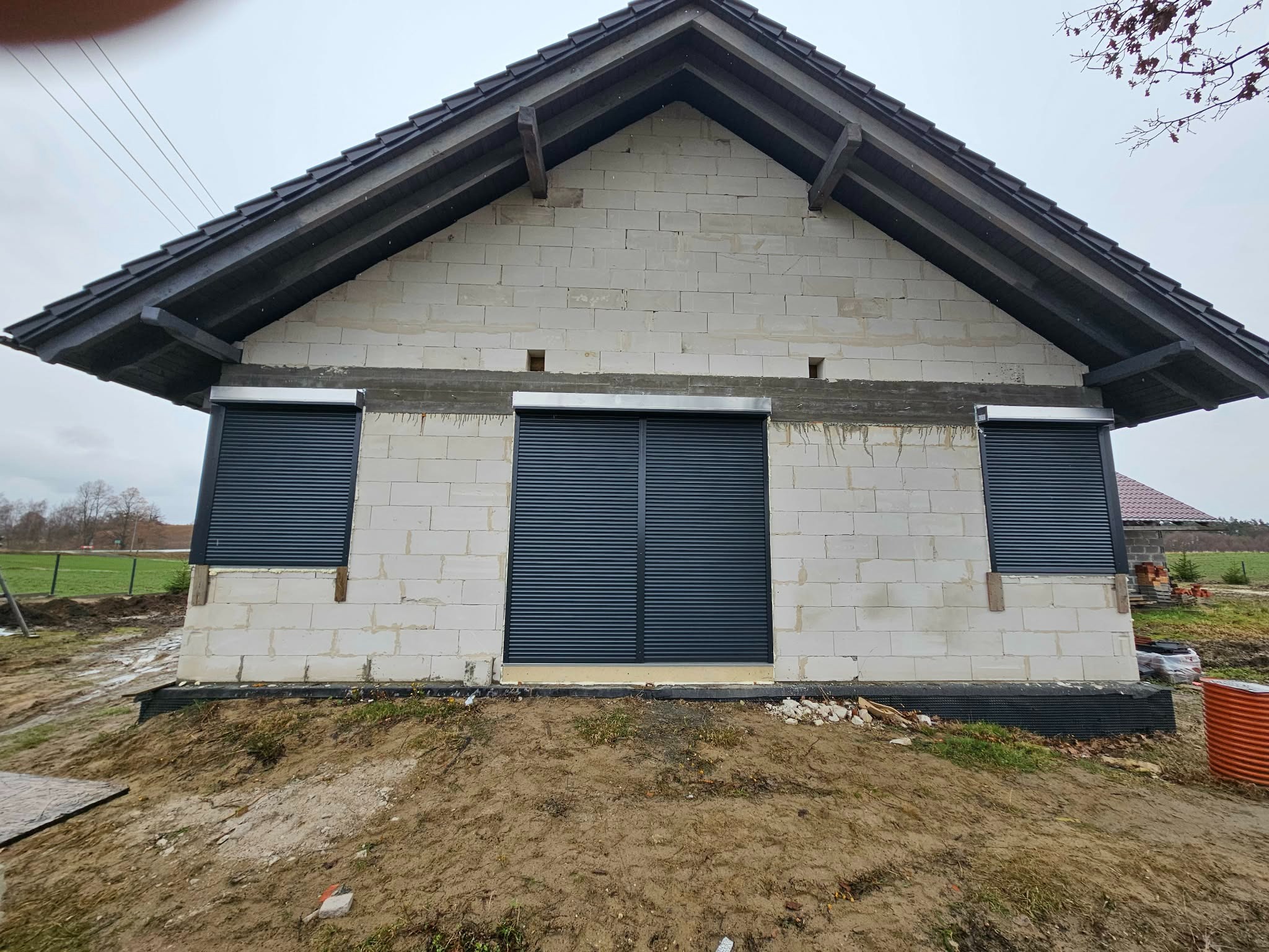 Surowe ściany budynku z trzema oknami zabezpieczonymi czarnymi roletami zewnętrznymi, widok z zewnątrz na tle pochmurnego nieba i częściowo zagospodarowanego terenu.