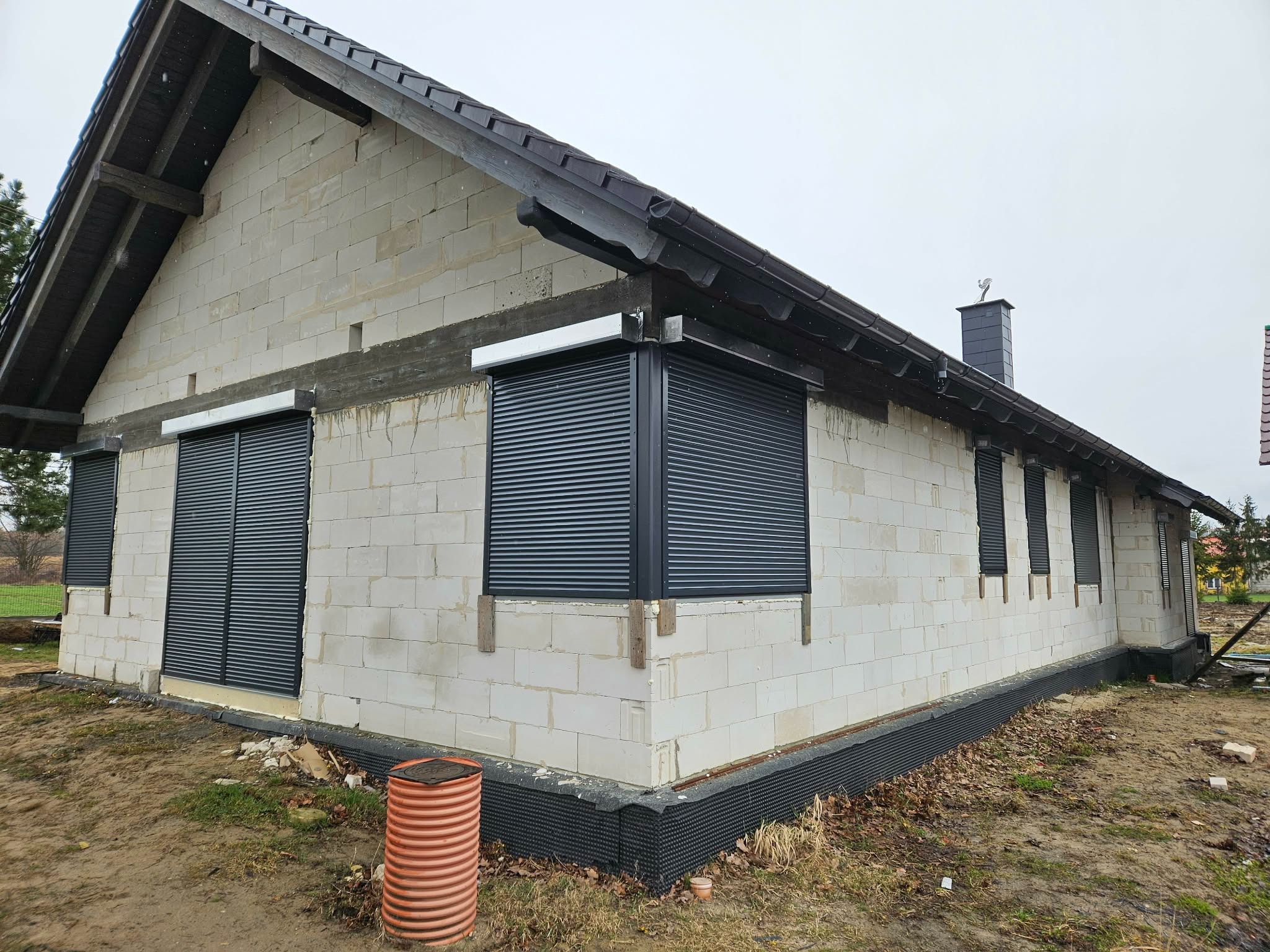 Dom w trakcie budowy z zamontowanymi, grafitowymi roletami zewnętrznymi na oknach. Widoczna surowa elewacja z betonu komórkowego i czarne orynnowanie.