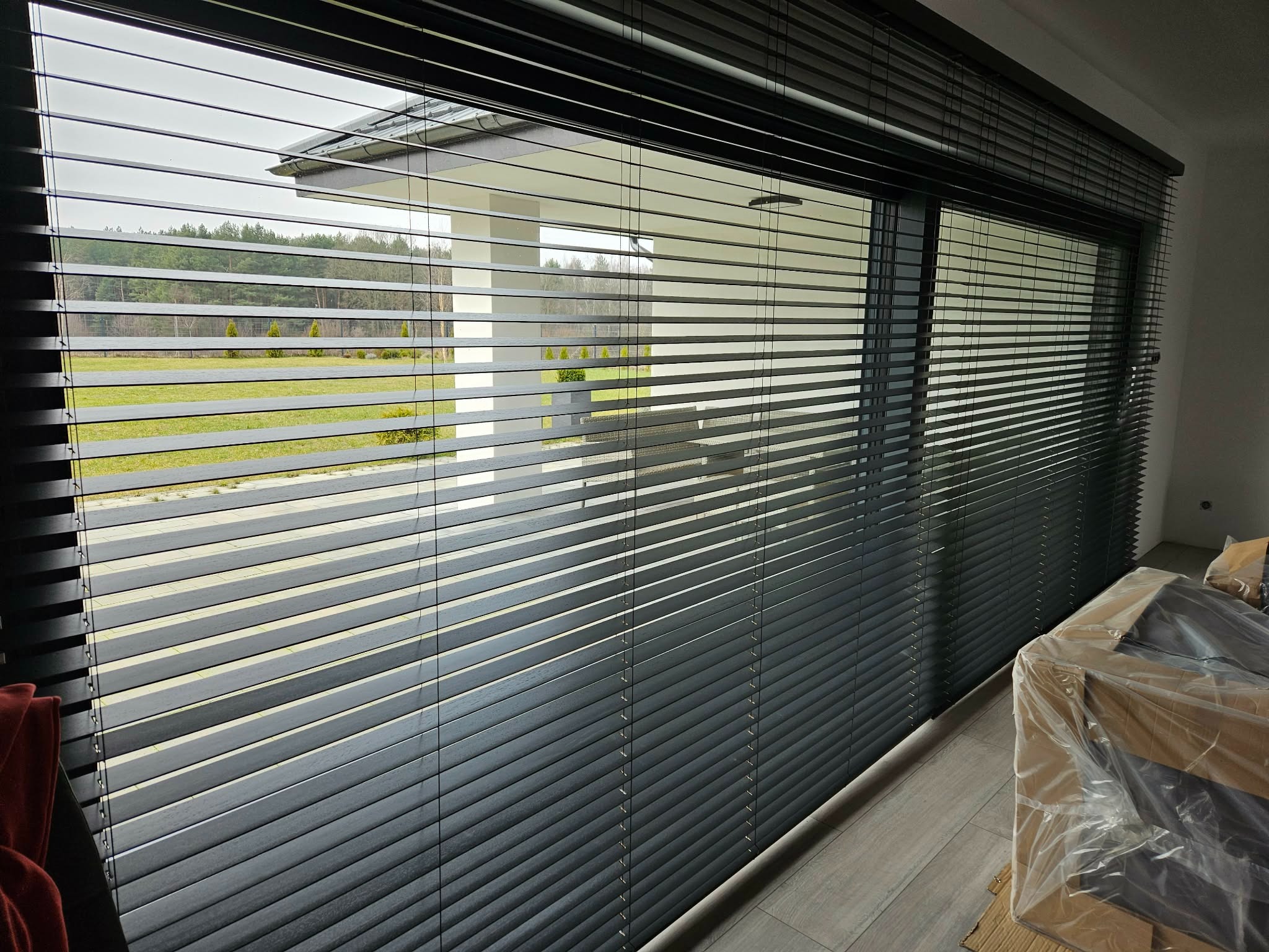 Ciemne żaluzje poziome zamontowane w oknie, widok na zewnątrz z zielenią i nowoczesną architekturą. Częściowo zasłonięte meble w folii ochronnej.