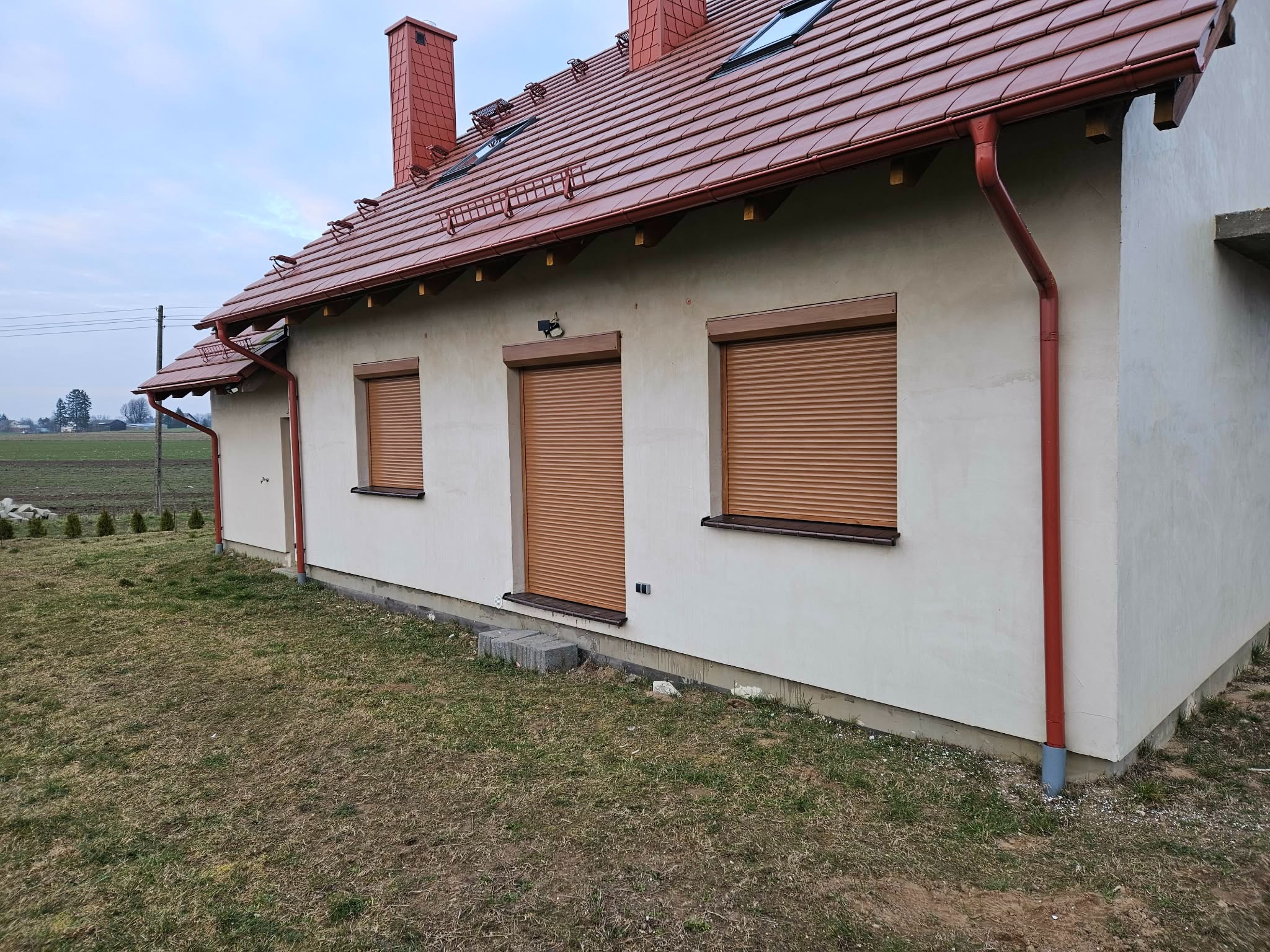 Dom jednorodzinny z brązowymi roletami zewnętrznymi na oknach i drzwiach, brązowe rynny i dachówka. Widok z boku na elewację budynku.