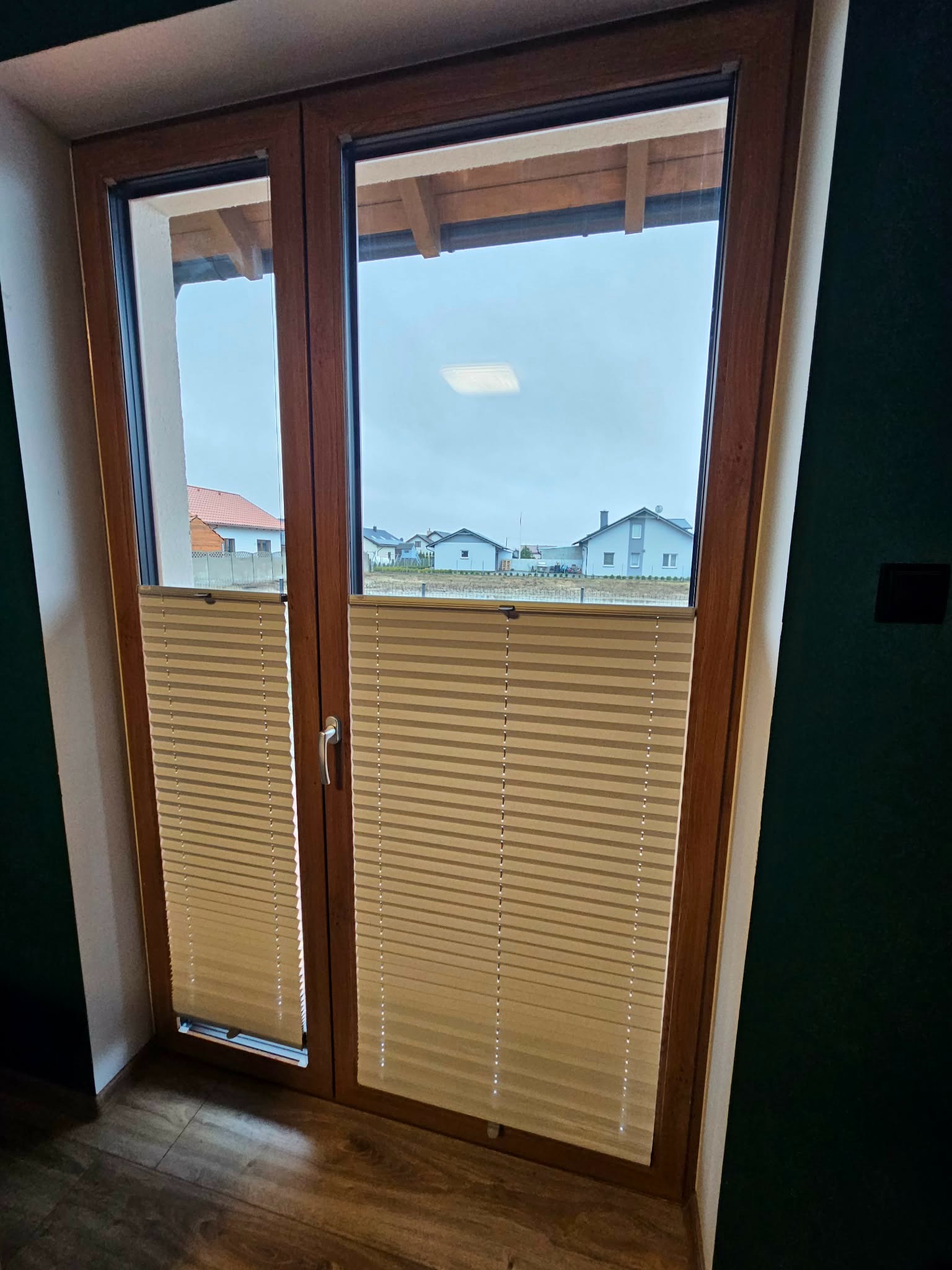 Drewniane okno z beżowymi plisowanymi roletami wewnętrznymi, widok na domy za oknem. Plisy częściowo zasłonięte, widoczna faktura materiału i mechanizm regulacji.