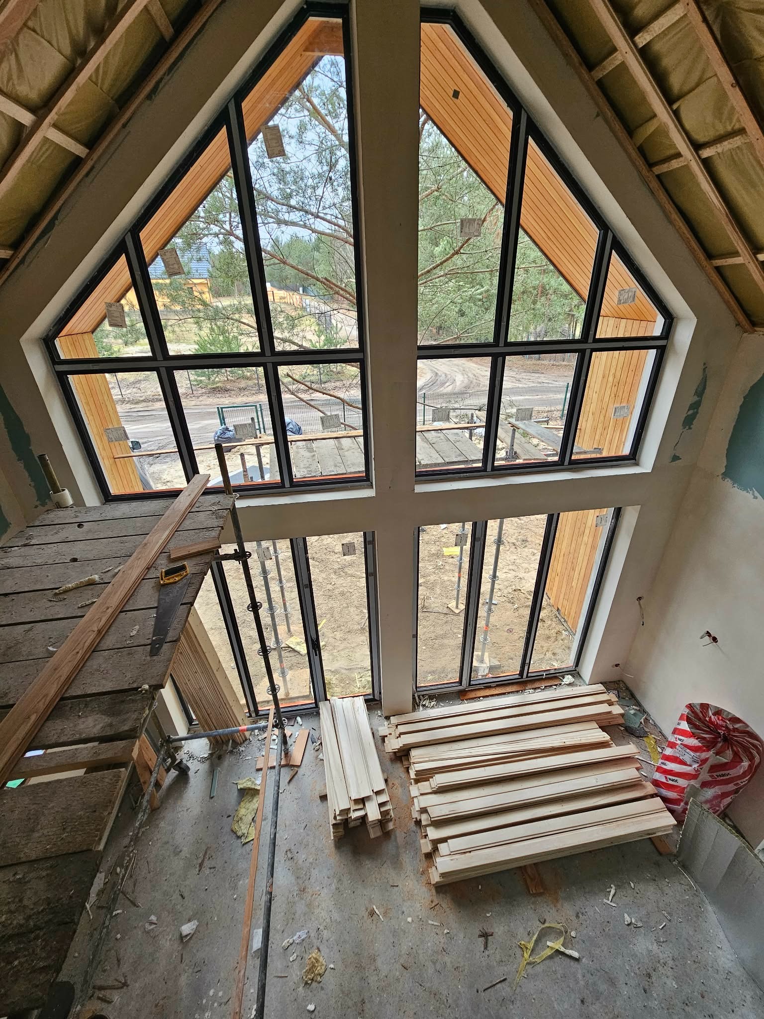 Montaż okien o nietypowym kształcie w surowym wnętrzu domu. Widoczne rusztowanie, deski i narzędzia. Okna z czarnymi ramami, widok na zewnątrz.