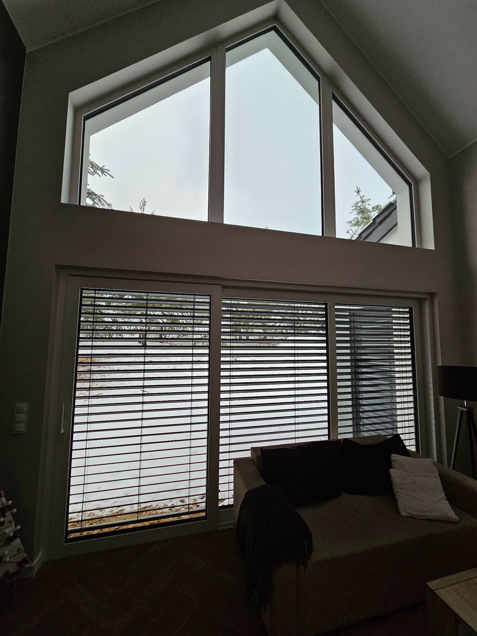 Wnętrze z dużym oknem z żaluzjami poziomymi i trójkątnym oknem powyżej, widok na zaśnieżony krajobraz. Minimalistyczny design, stonowana kolorystyka.