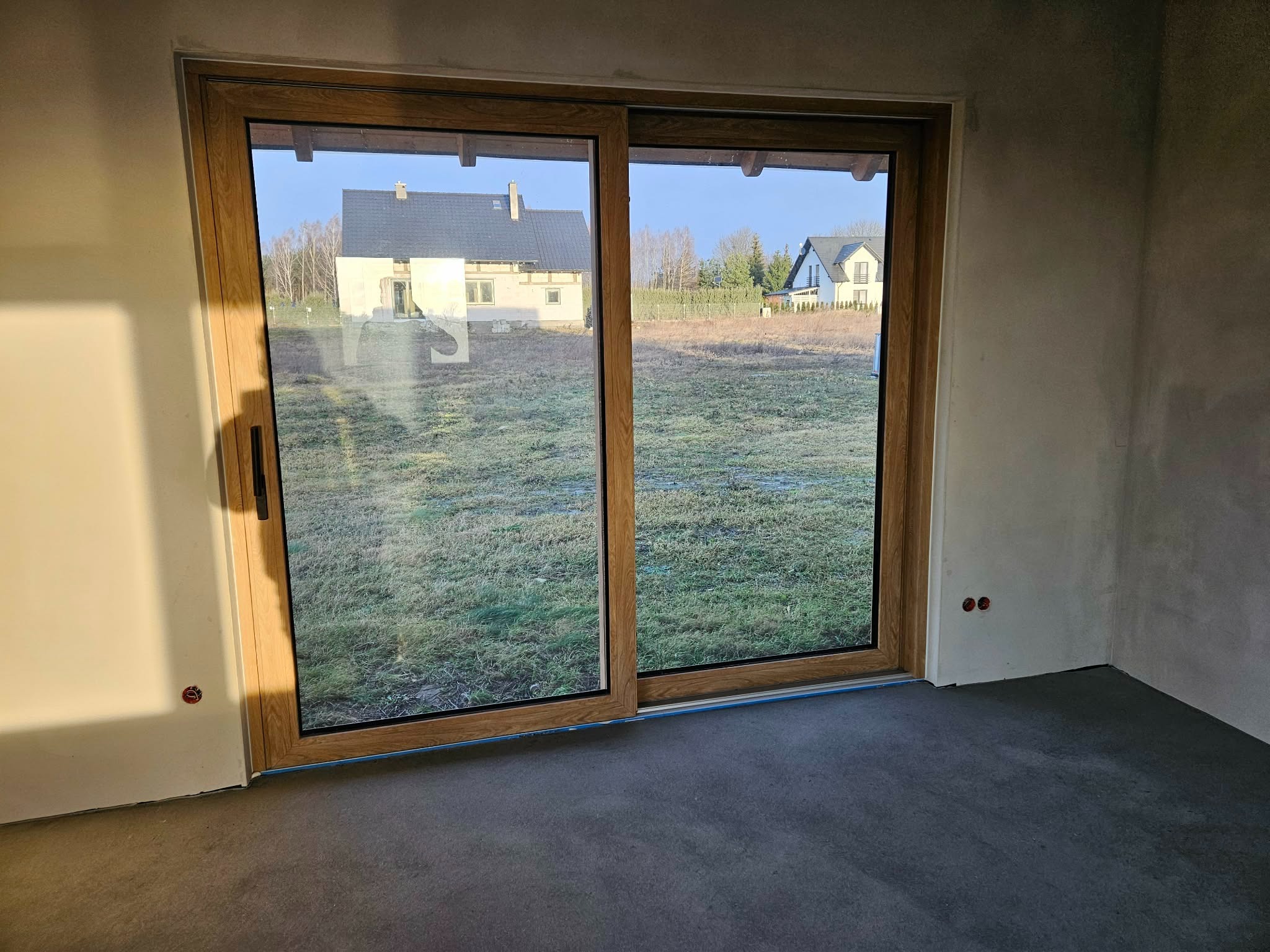 Przesuwne okno tarasowe z drewnianą ramą, widok na zewnątrz z dwoma domami w tle. Surowe ściany wewnątrz, wylewka podłogowa.