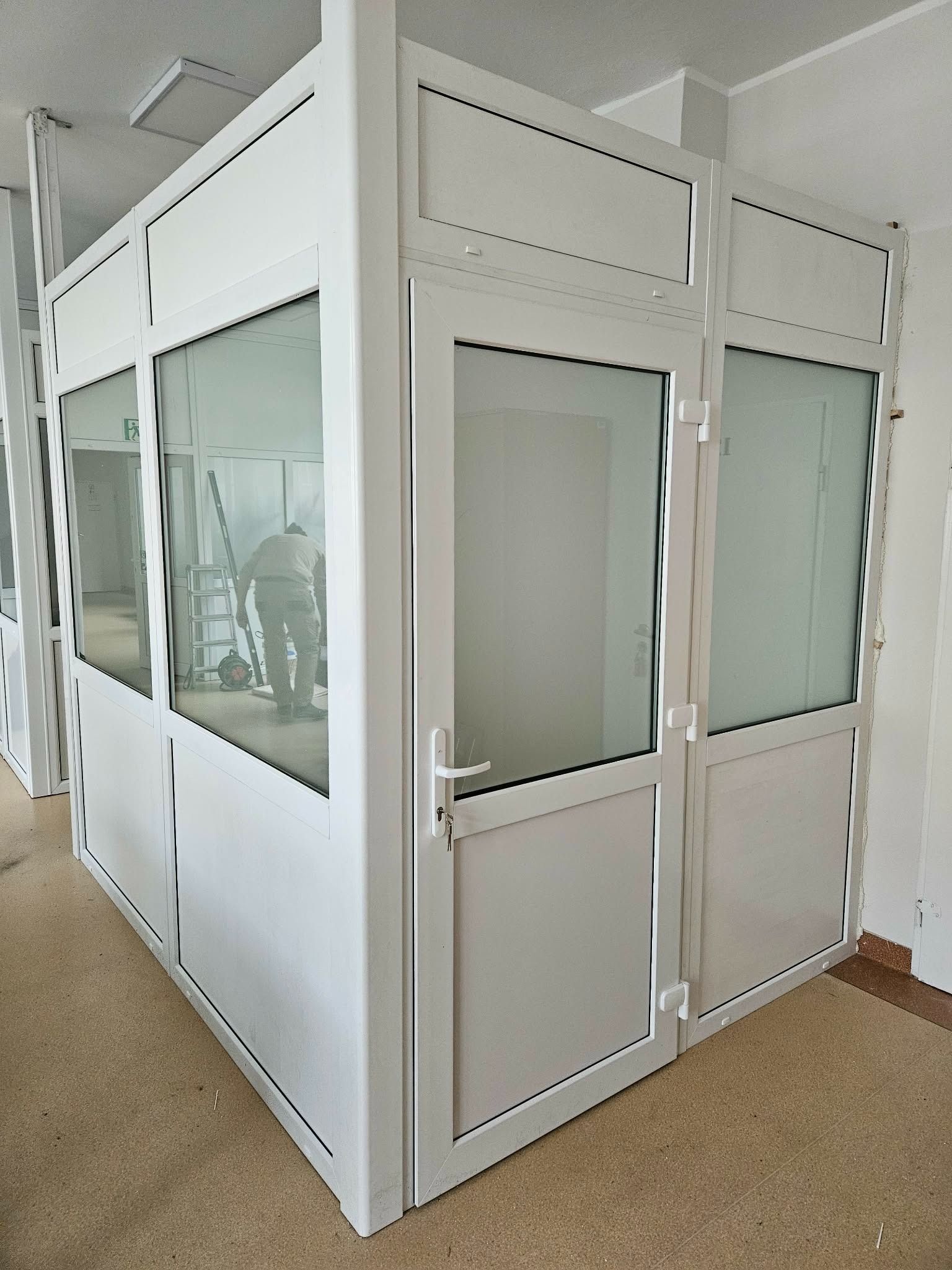 Biała, przeszklona konstrukcja z drzwiami, prawdopodobnie biurowa, z widoczną osobą pracującą wewnątrz. Widoczne detale wykończenia i oświetlenie.