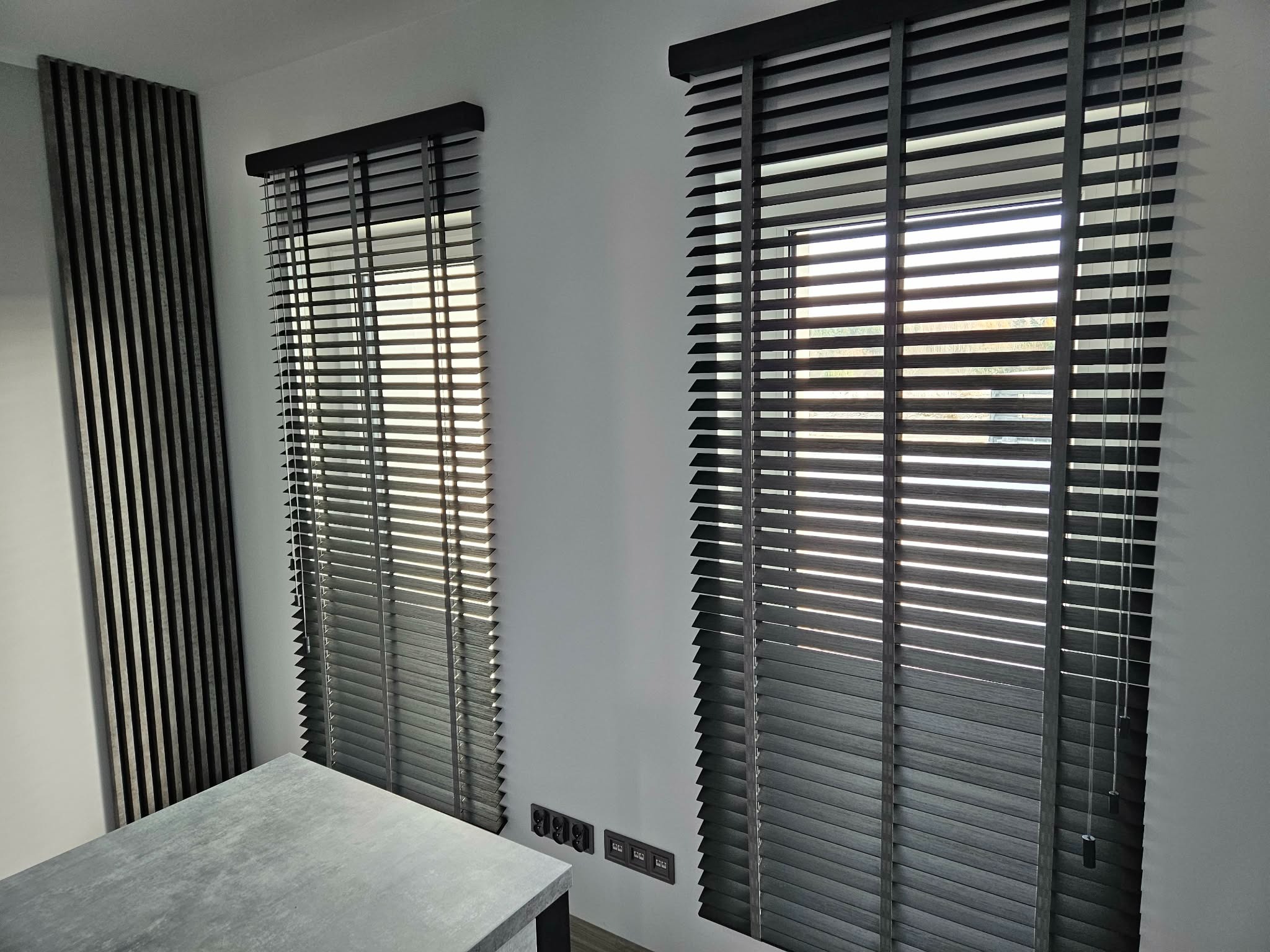Dwa okna z drewnianymi żaluzjami w odcieniach szarości, z widocznymi sznurkami do regulacji. Obok dekoracyjny panel ścienny z pionowych listew w podobnej kolorystyce.