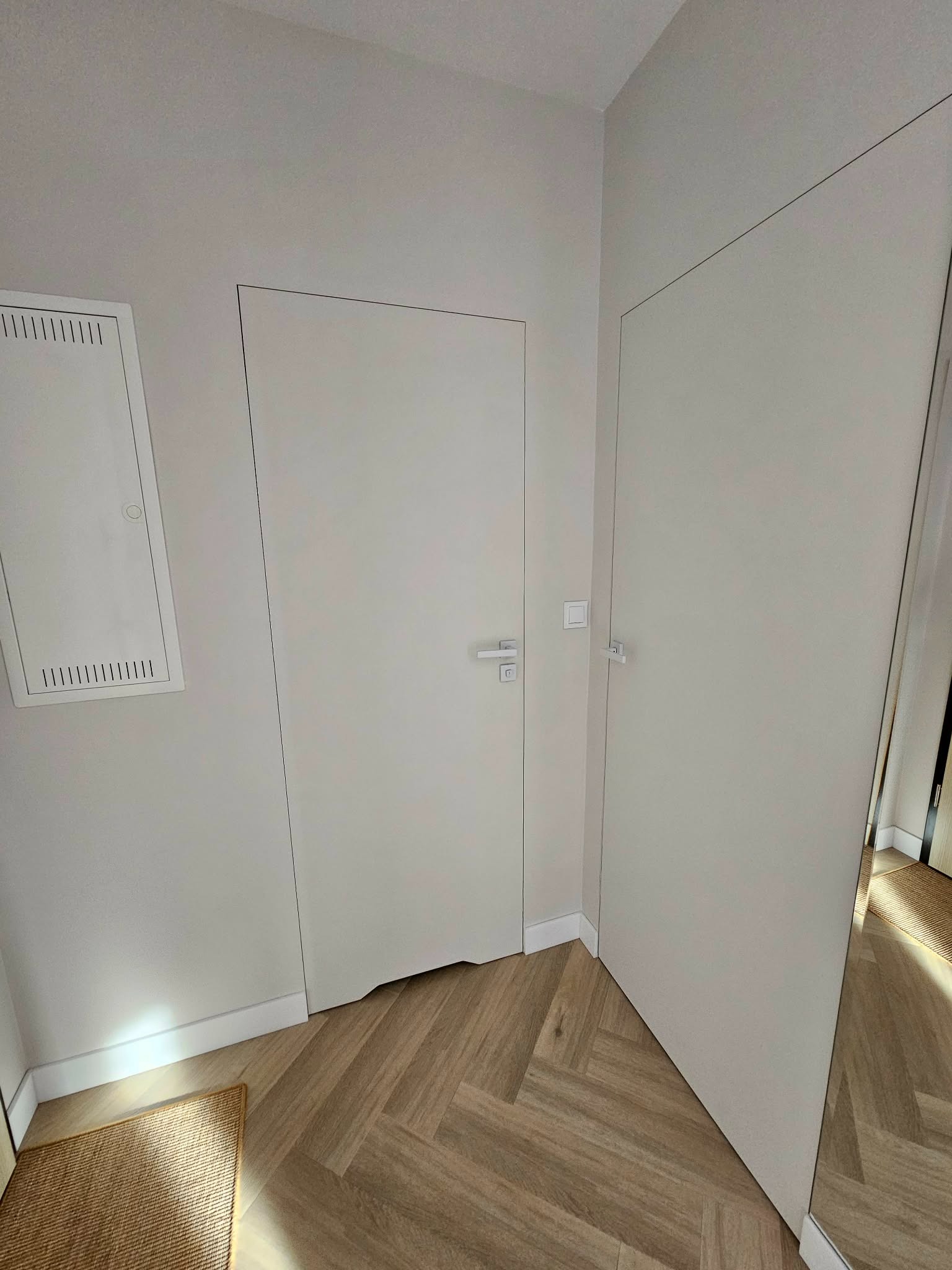 Minimalistyczne, białe drzwi wewnętrzne z ukrytą ościeżnicą, wkomponowane w jasne wnętrze z parkietem w jodełkę. Widoczny fragment dywanika i skrzynka elektryczna.
