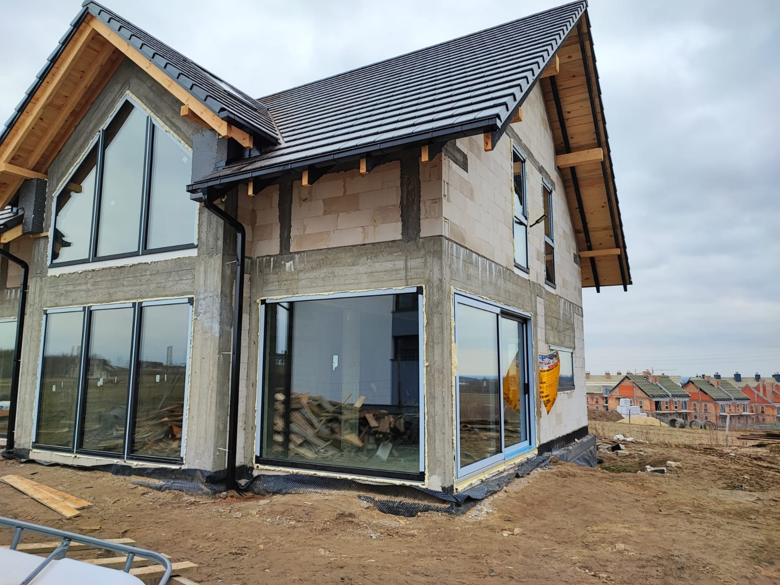 Dom w trakcie budowy z zamontowanymi dużymi oknami przesuwnymi i trójkątnym oknem na szczycie, widoczne materiały budowlane i inne domy w tle.