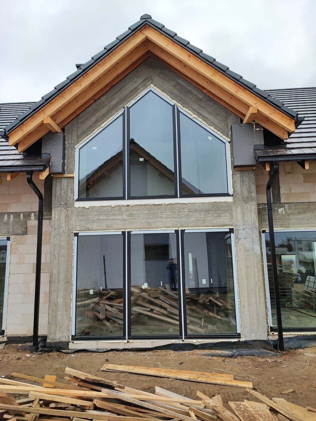 Surowy stan budynku z zamontowanymi oknami o czarnych ramach, widoczne drewniane elementy konstrukcji dachu oraz porozrzucane deski na zewnątrz.