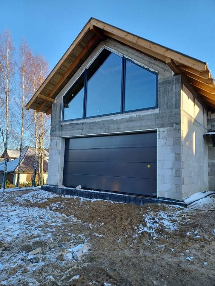 Dom w trakcie budowy z zamontowaną ciemnoszarą bramą garażową segmentową i dużym trójkątnym oknem z czarną ramą na szczycie, widoczne bloczki betonowe i ośnieżona ziemia.