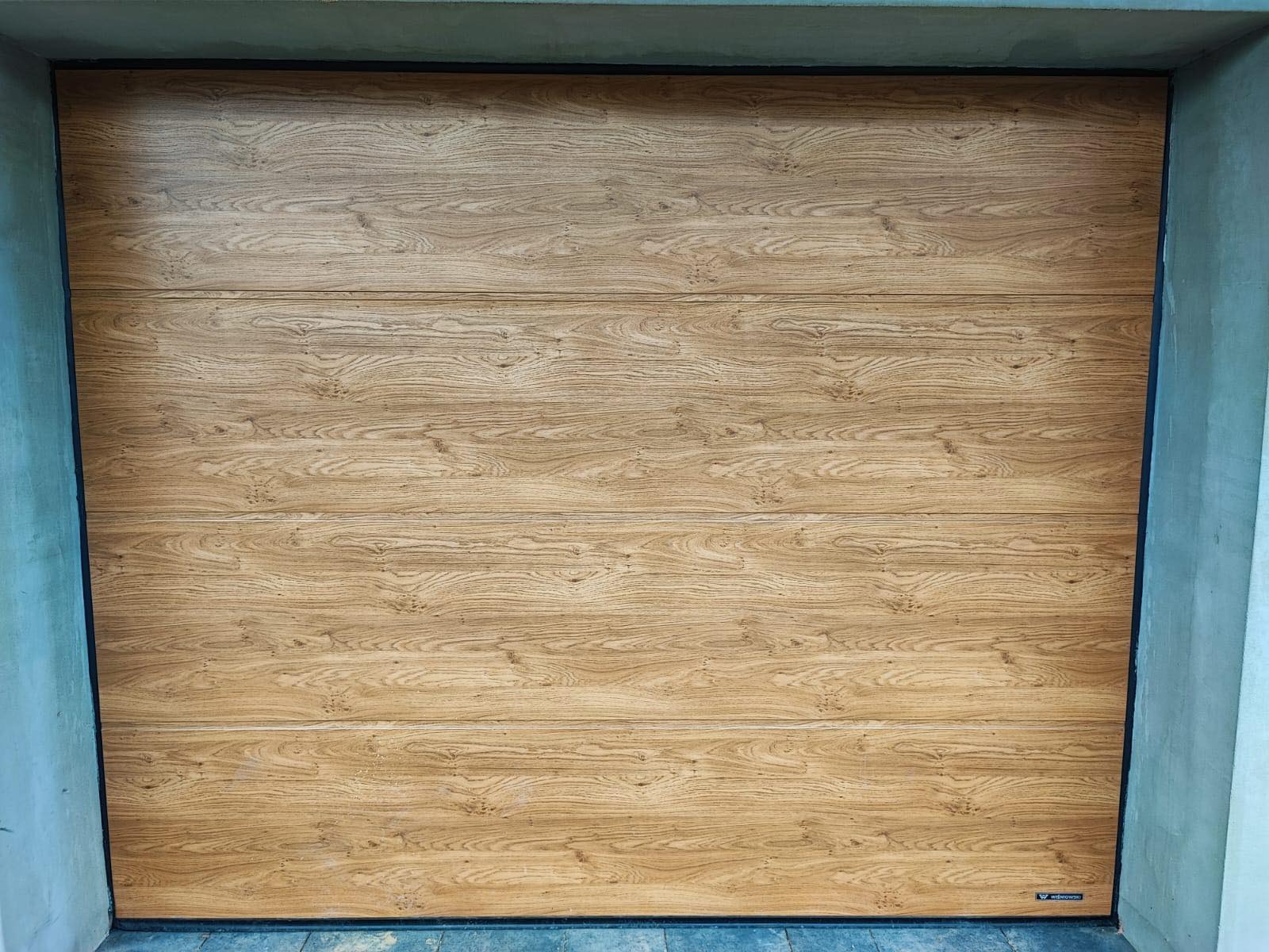 Zamknięta brama garażowa segmentowa z paneli imitujących jasne drewno dębowe, widoczna od wewnątrz, z czarną ramą i logo producenta w prawym dolnym rogu.