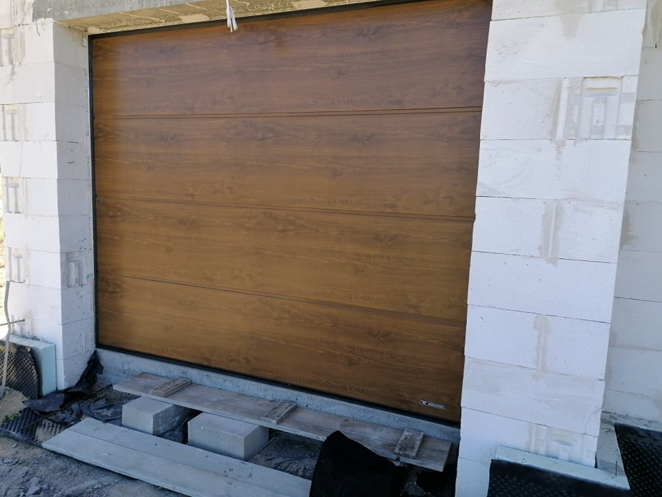Zamknięta brama garażowa segmentowa w kolorze drewna, zamontowana w surowym otworze z białych bloczków betonowych, widoczne podpory montażowe.