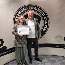 Dwie osoby, kobieta z dredami i mężczyzna, stoją przed ścianką z logo Omni Hypnosis Training Center, trzymając certyfikat ukończenia kursu.