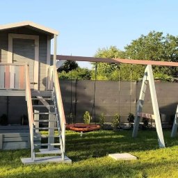 Krzysztof Majewski - Drewniany plac zabaw z domkiem na podeście, schodami i huśtawką typu bocianie gniazdo, pomalowany na szaro z różowymi akcentami, na tle zielonego trawnika i ciemnego ogrodzenia.