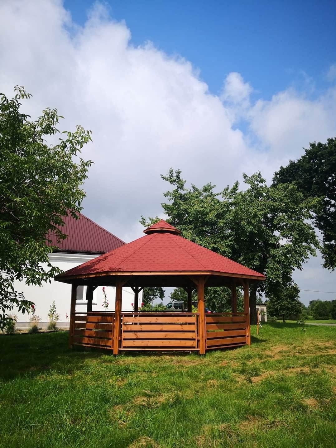 Ośmiokątna altana ogrodowa z czerwonym dachem i drewnianą balustradą, otoczona zielonym trawnikiem i drzewami, w tle widoczny budynek z czerwonym dachem.