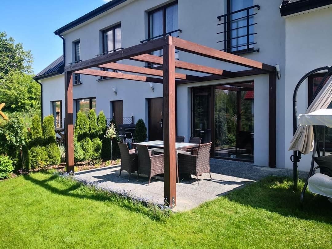 Drewniana konstrukcja pergoli z ciemnego drewna nad betonowym tarasem z meblami ogrodowymi, przylegająca do białego domu z dużymi oknami i zadbanym trawnikiem.