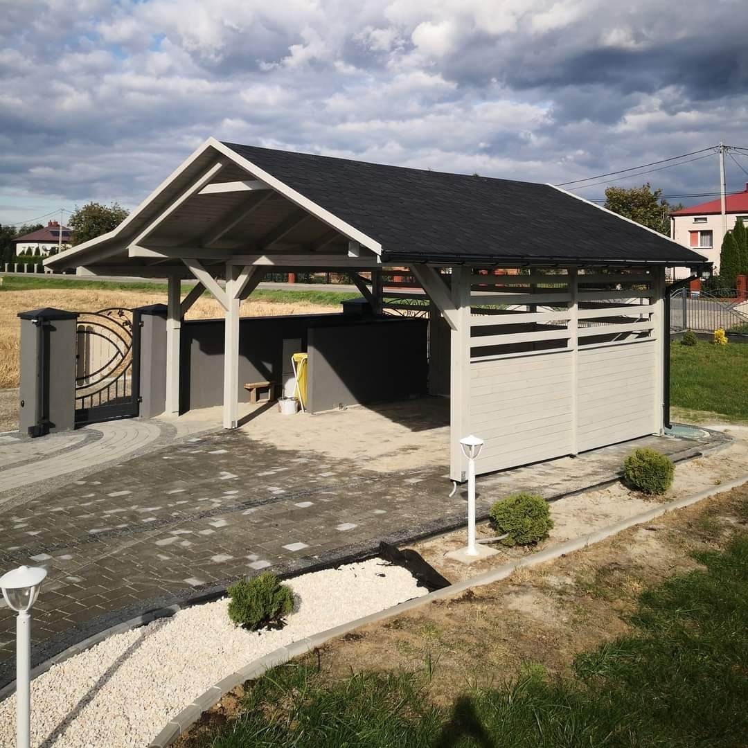 Zadaszona wiata garażowa z drewnianą konstrukcją w odcieniach szarości, z czarnym dachem, przylegająca do szarego ogrodzenia z ozdobną bramą, oświetlona lampami ogrodowymi i otoczona kostką brukową...