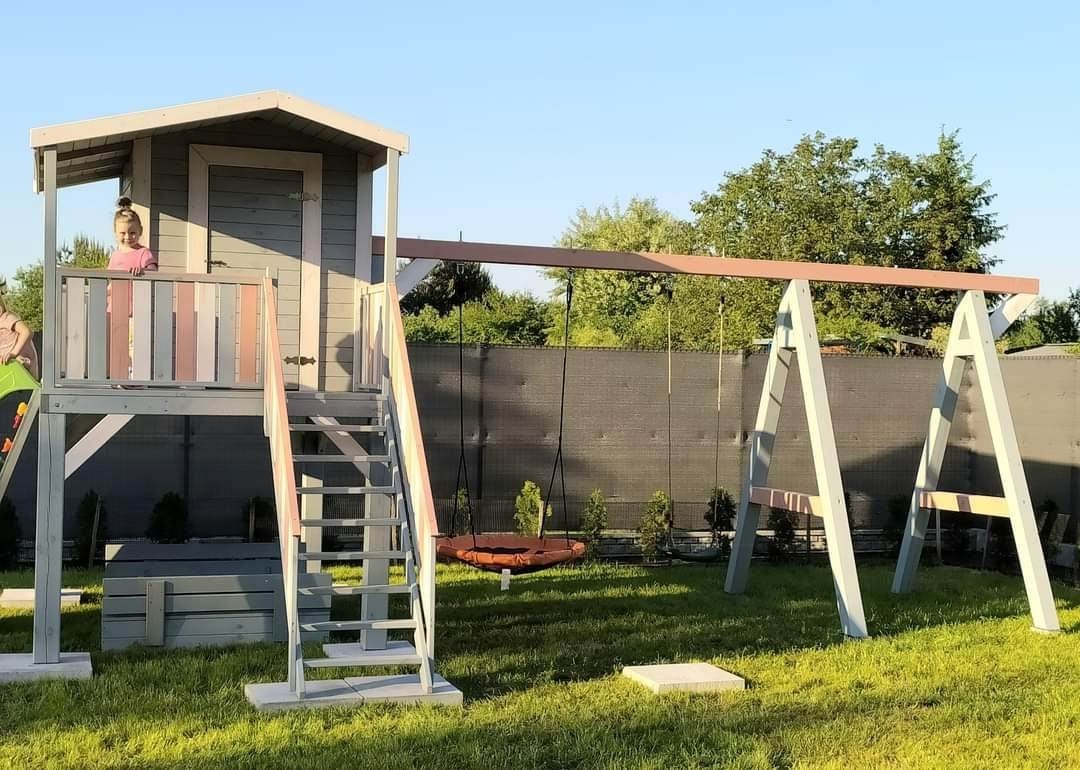 Drewniany plac zabaw z domkiem na podeście, schodami i huśtawką typu bocianie gniazdo, pomalowany na szaro z różowymi akcentami, na tle zielonego trawnika i ciemnego ogrodzenia.