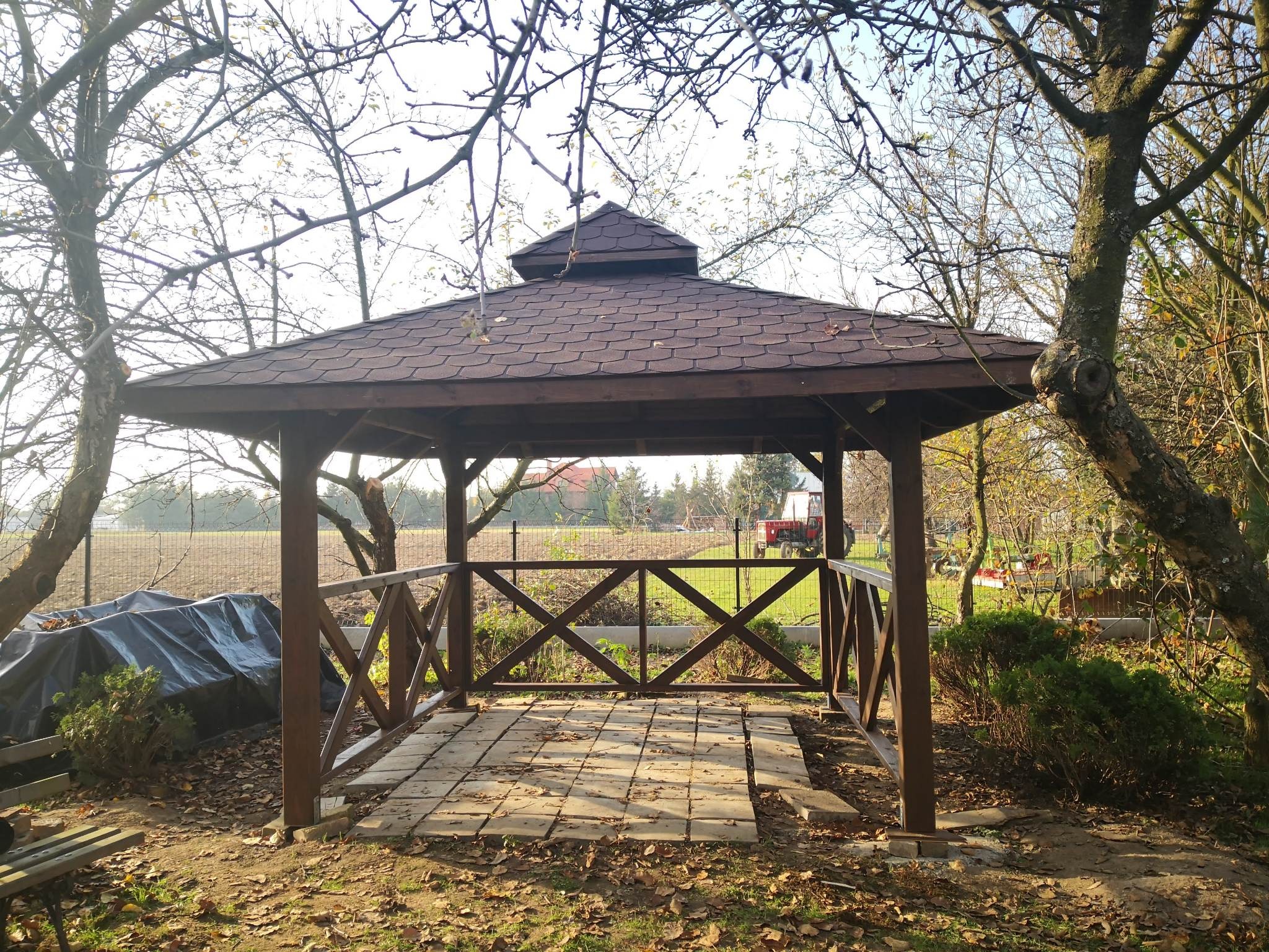 Drewniana altana z czterospadowym dachem z gontu bitumicznego, otoczona jesiennym ogrodem z widocznym traktorem w tle.