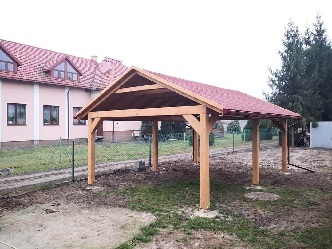 Drewniana konstrukcja z czerwonym dachem, prawdopodobnie wiata garażowa, na tle domu jednorodzinnego i ogrodzenia z siatki.