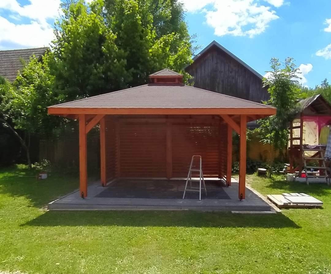 Drewniana altana z brązowym dachem na betonowej podstawie, z drabinką wewnątrz, umieszczona w ogrodzie z zielonym trawnikiem i zabudowaniami w tle.
