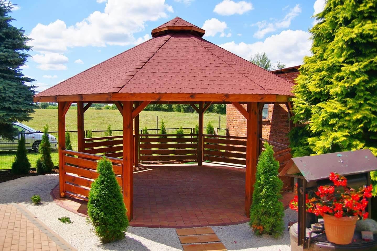 Ośmiokątna drewniana konstrukcja z czerwoną dachówką, otoczona białym żwirem i niskimi tujami, z widokiem na zielone pole w tle.