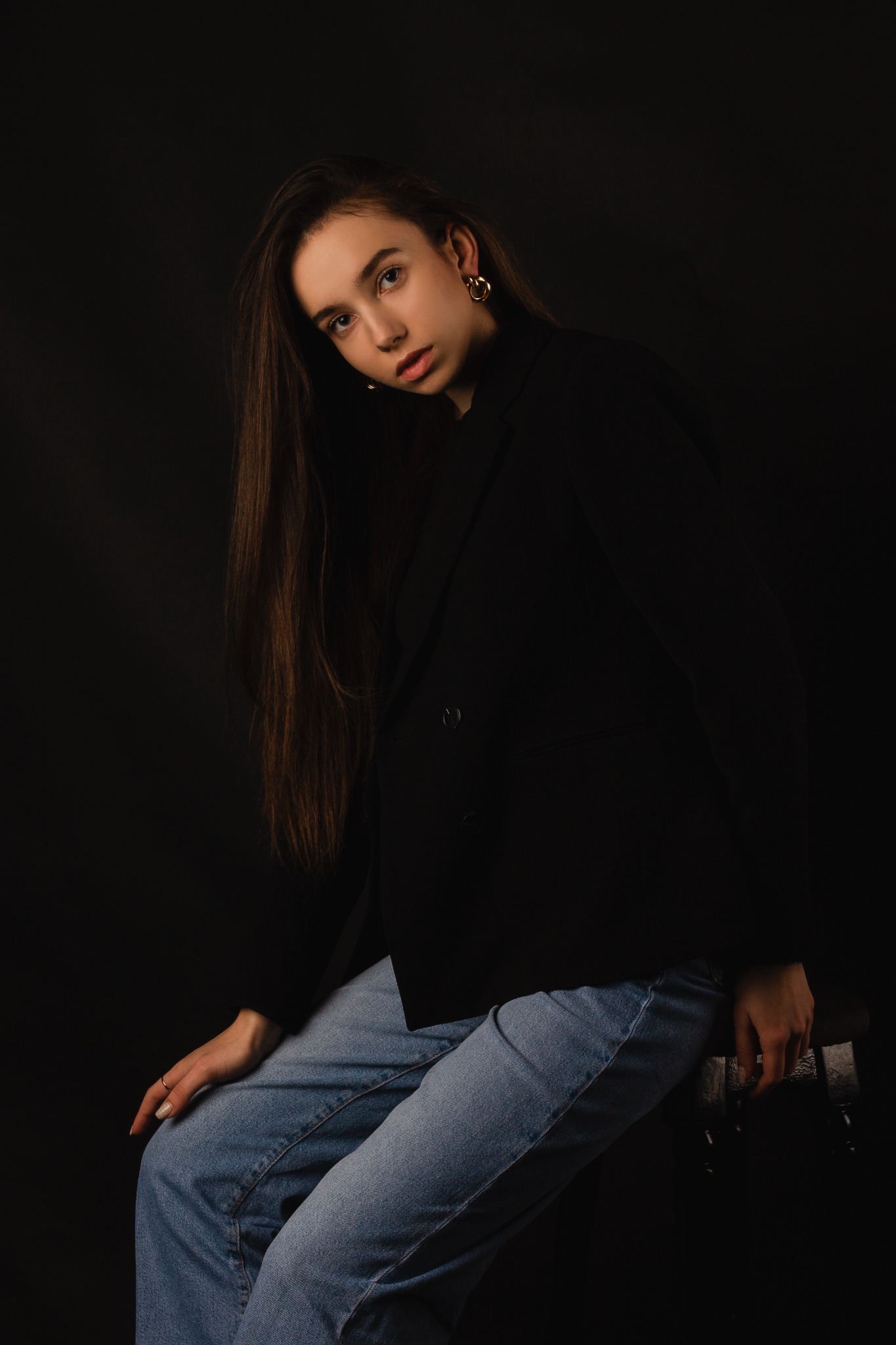 Portret młodej kobiety w czarnej marynarce i jeansach, siedzącej na stołku na ciemnym tle, z długimi brązowymi włosami i złotymi kolczykami.