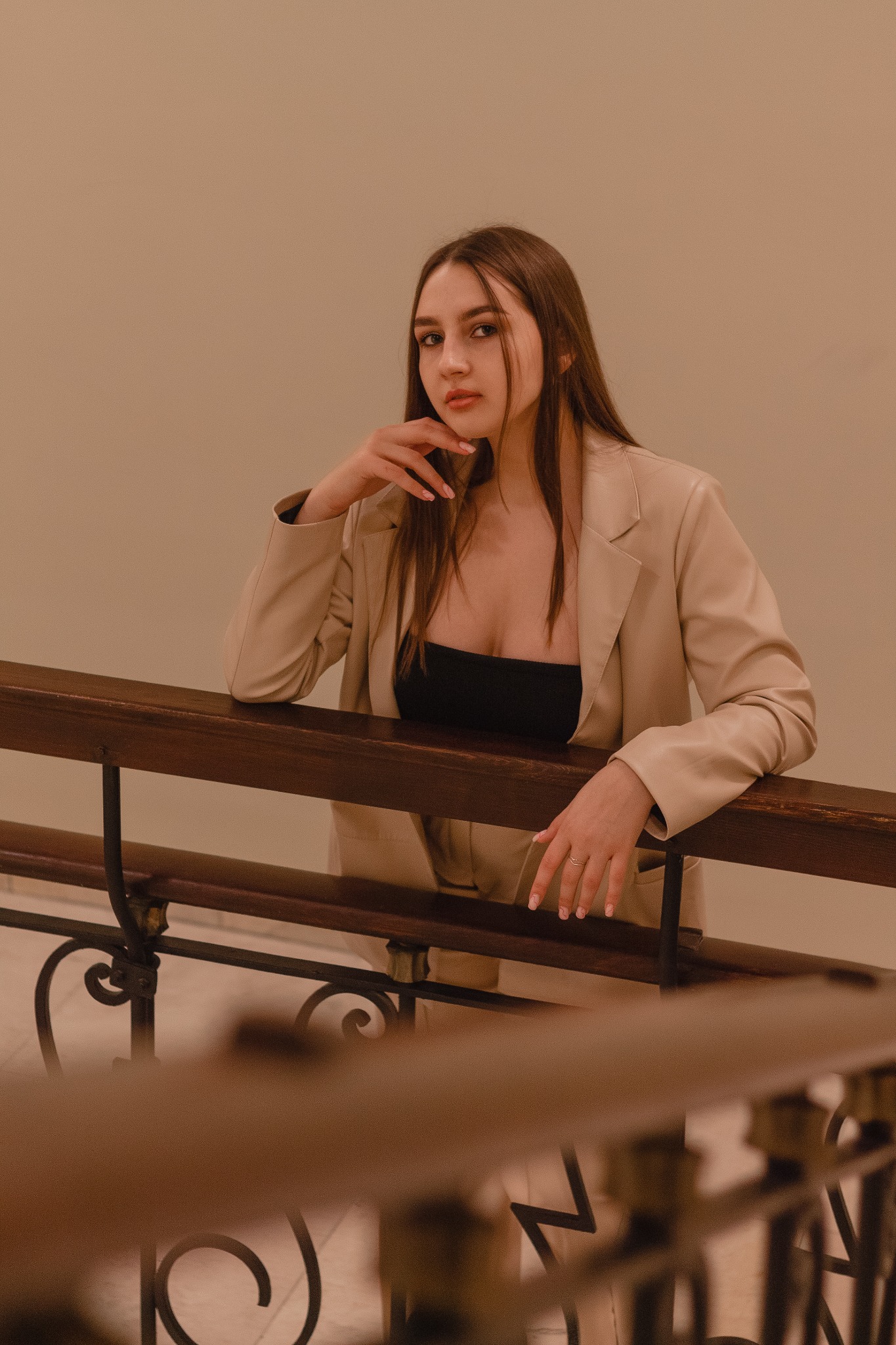 Portret kobiety w beżowym garniturze opierającej się o drewnianą balustradę schodów z metalowymi zdobieniami.