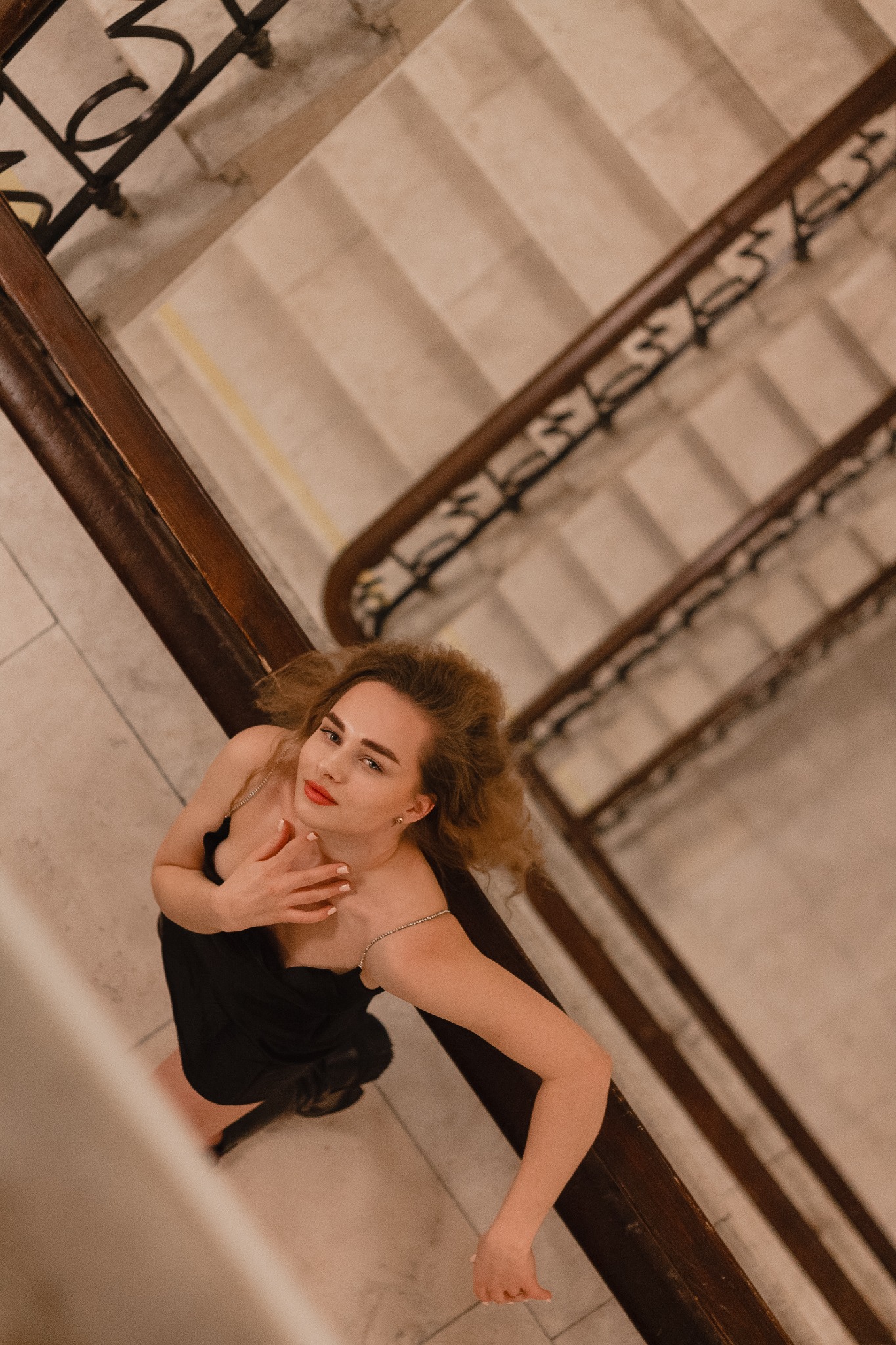 Kobieta w czarnej sukience pozuje na zabytkowej klatce schodowej, widok z góry, oświetlenie podkreśla kręcone włosy i czerwone usta.