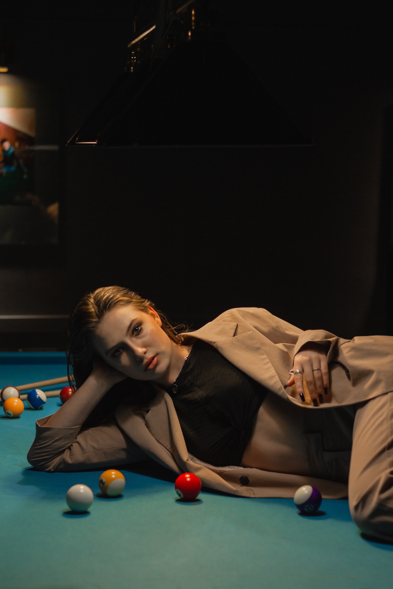 Kobieta w beżowym garniturze leży na stole bilardowym z kolorowymi bilami, spoglądając w obiektyw, oświetlona punktowym światłem.
