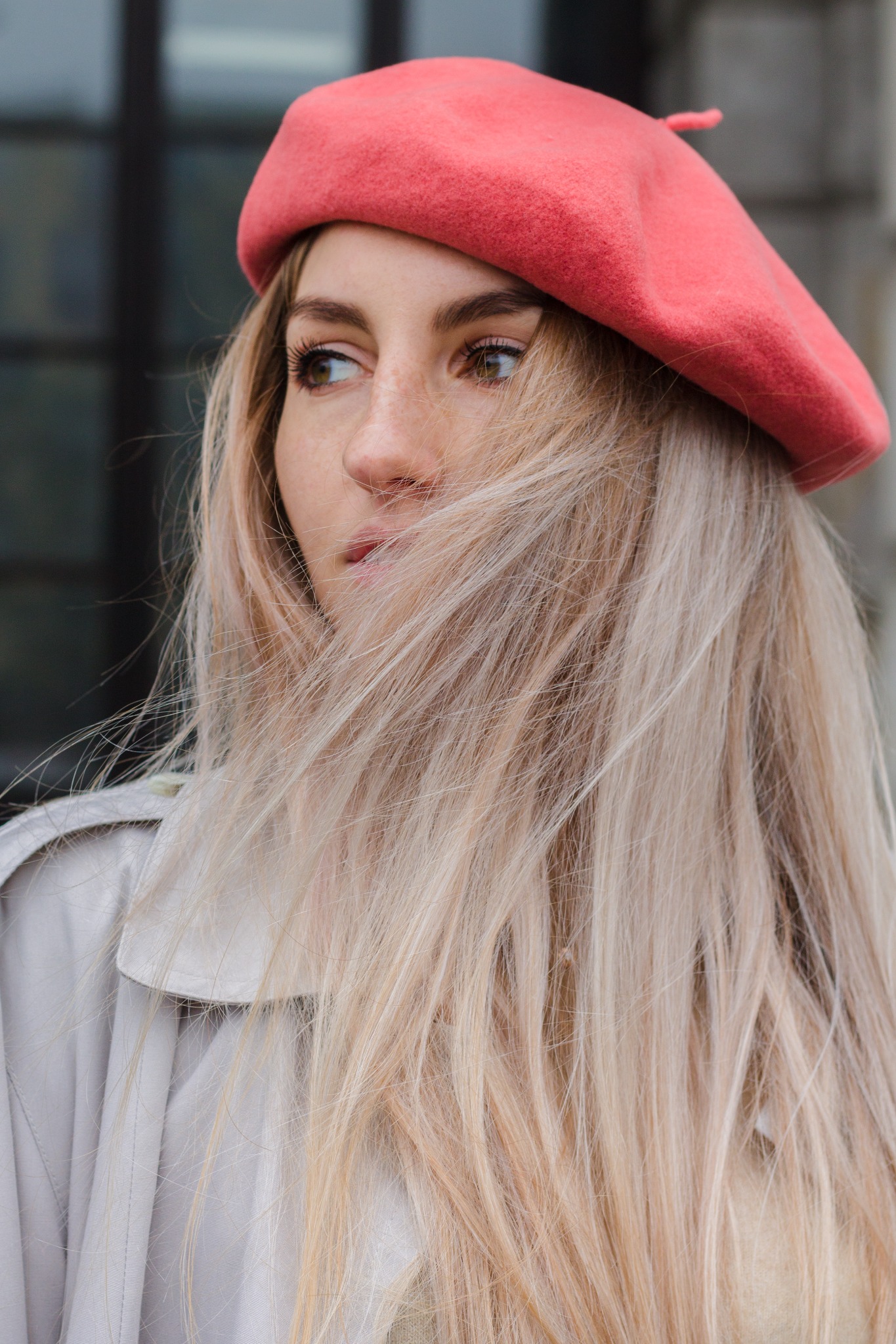 Portret kobiety w czerwonym berecie, z blond włosami przesłaniającymi część twarzy, w jasnym płaszczu, zamyślona, na tle budynku.