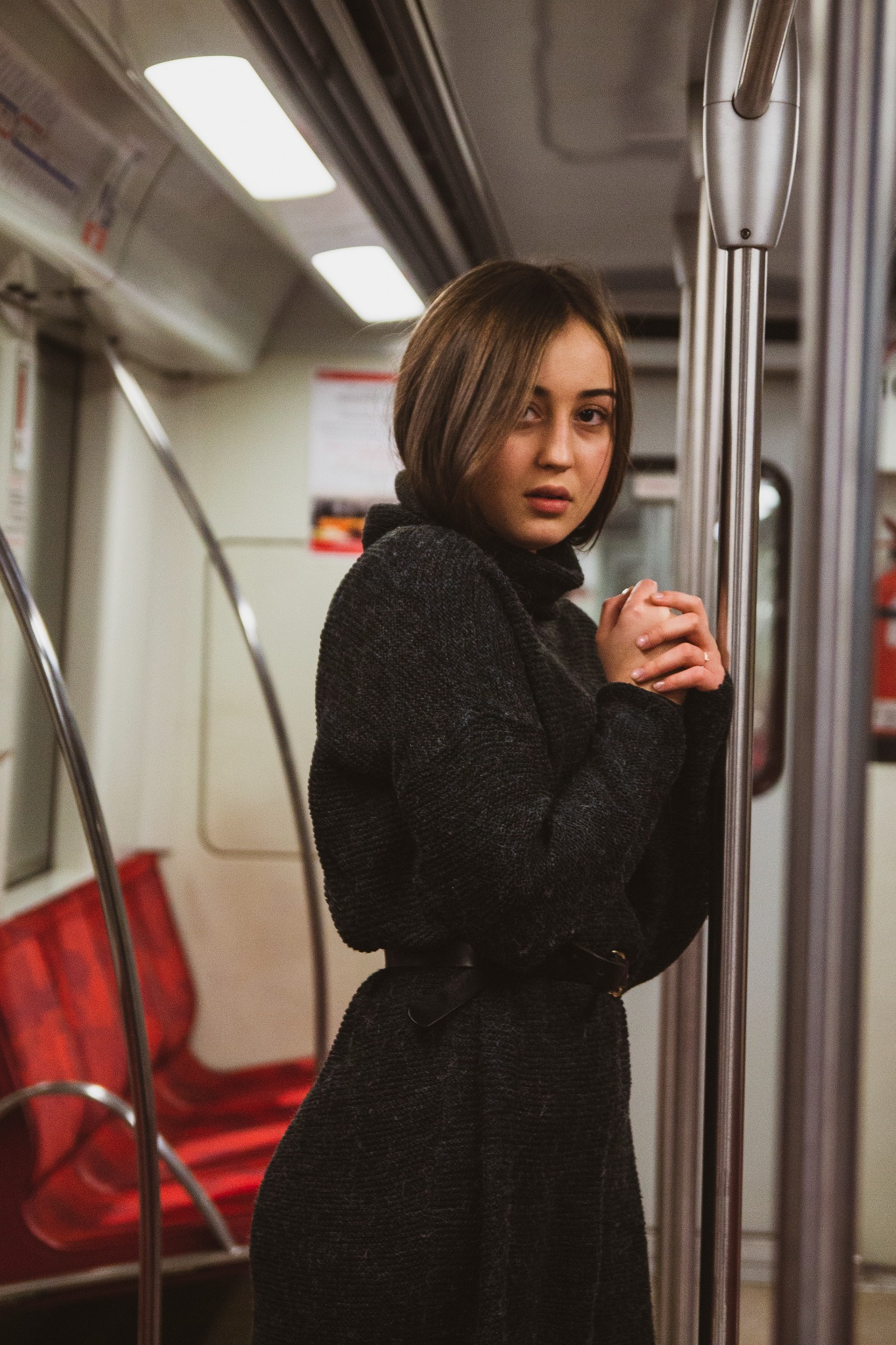 Portret kobiety w ciemnym swetrze z golfem, opierającej się o metalowy drążek w wagonie metra, z zaciśniętymi dłońmi i zamyślonym spojrzeniem.