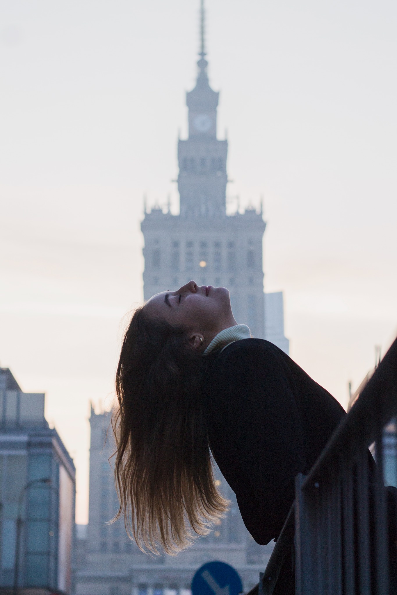Kobieta w czarnym płaszczu odchylona do tyłu z zamkniętymi oczami na tle Pałacu Kultury w Warszawie, ujęcie z dołu.