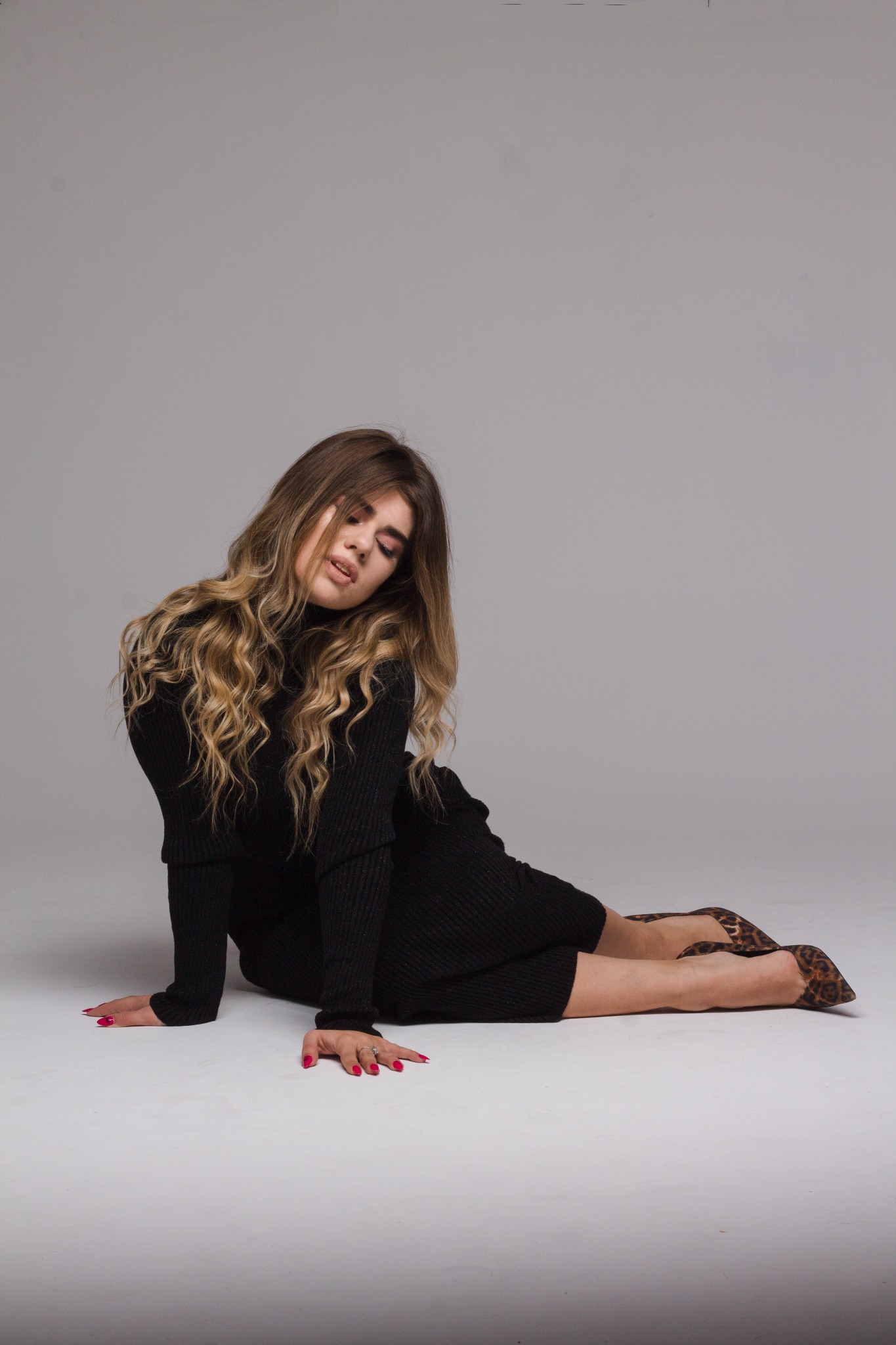 Kobieta w czarnej sukience z golfem i butach w panterkę pozuje siedząc na podłodze w studio, z delikatnym makijażem i rozpuszczonymi blond włosami.