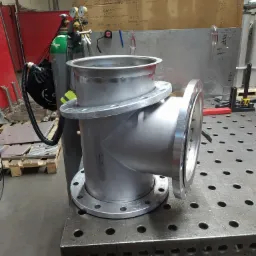 Metalowy element konstrukcyjny z kołnierzami, prawdopodobnie rura, leżący na stole spawalniczym z otworami, w tle butle z gazem i karton z notatkami.