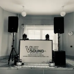 Czarno-białe zdjęcie sceny z nagłośnieniem i oświetleniem firmy Vast Sound, z dwoma kolumnami głośnikowymi JBL na statywach, dwoma reflektorami i podświetlanym panelem z logo firmy, dodatkowo...