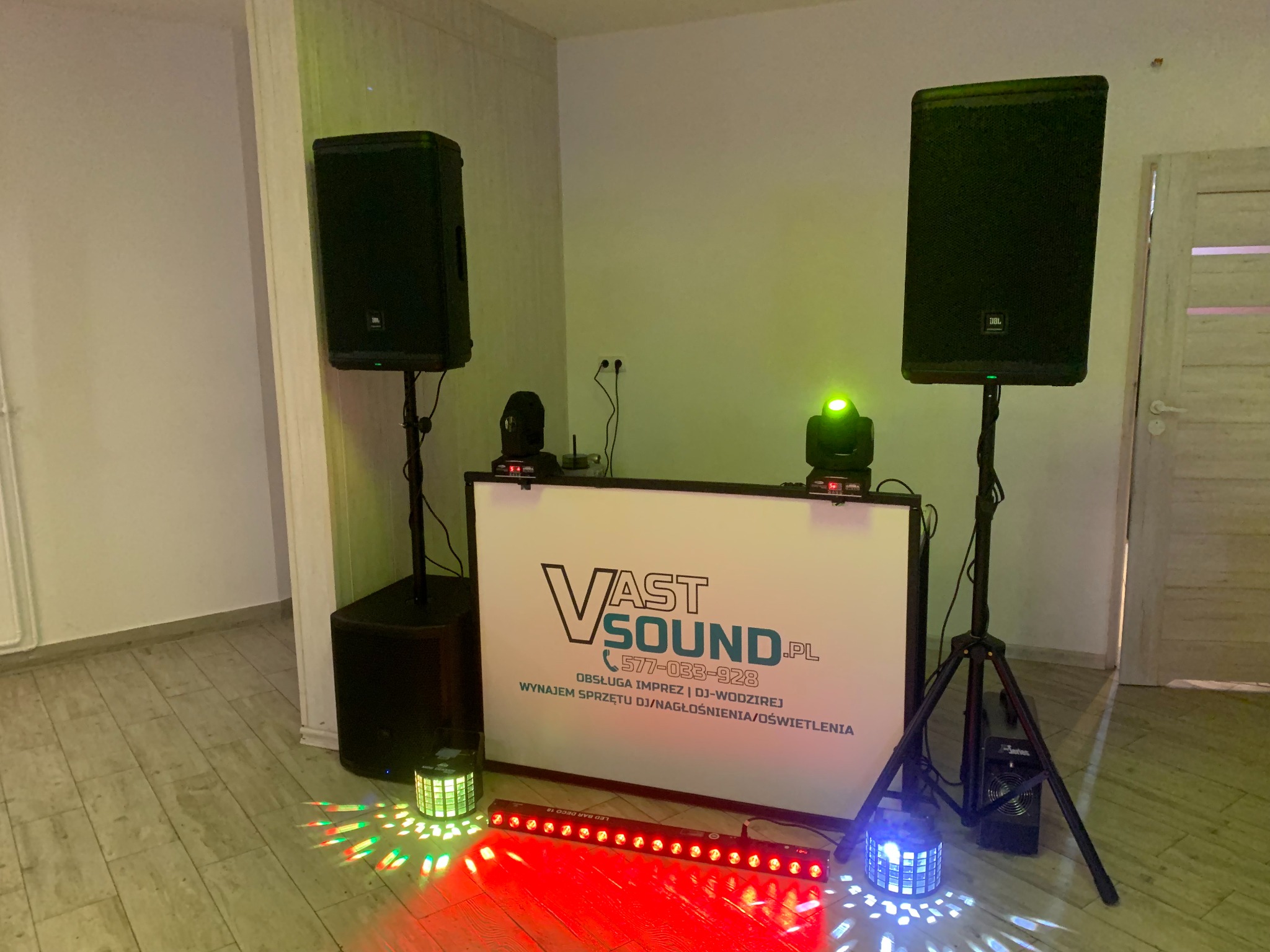 Sprzęt DJ-ski VastSound: dwa głośniki JBL na statywach, subwoofer, panel DJ-ski z logo firmy, reflektory oświetleniowe na podłodze rzucające kolorowe światła, przygotowany do imprezy w pomieszczeniu.