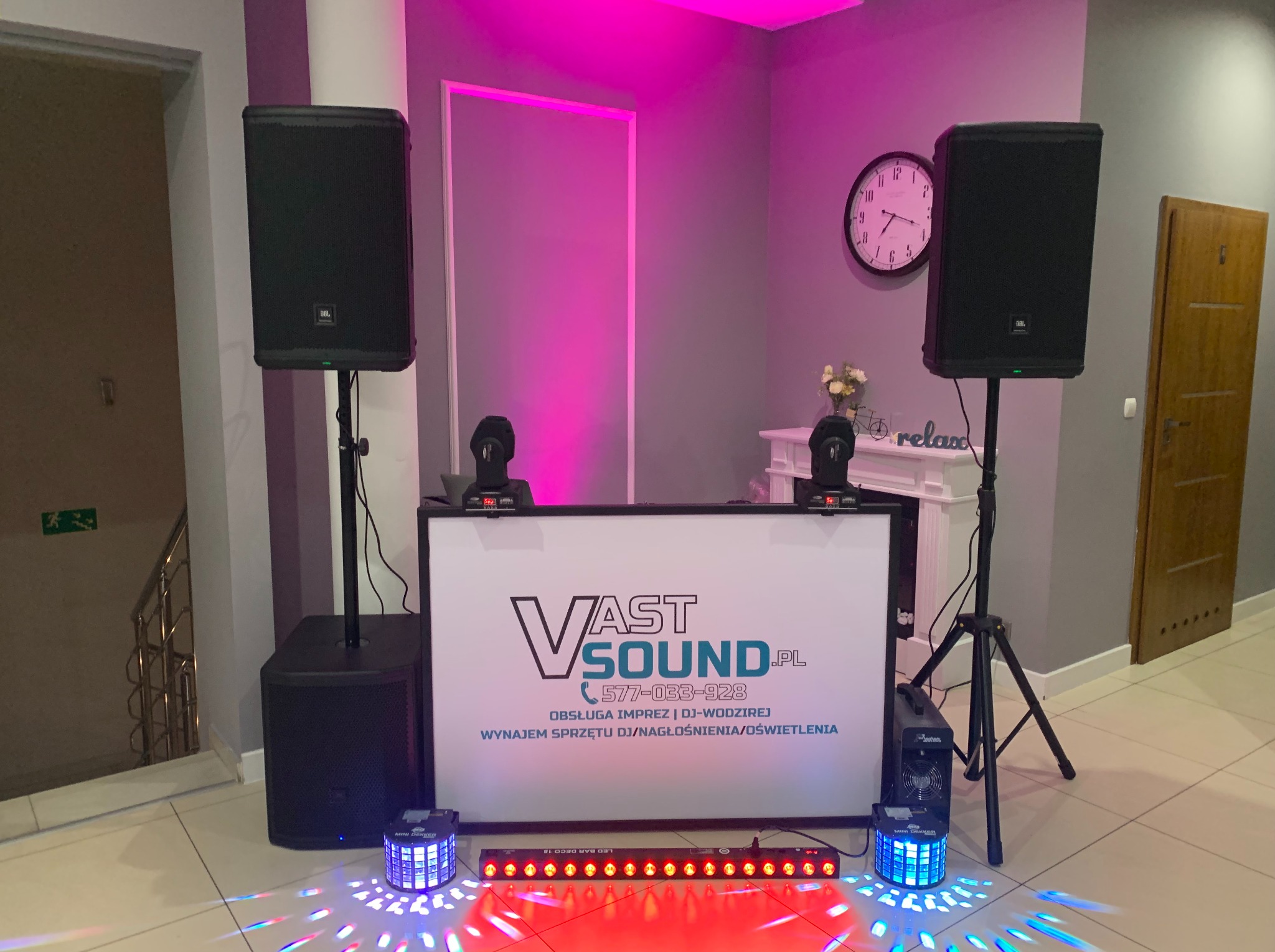 Sprzęt DJ-ski i oświetleniowy VastSound.pl ustawiony na sali: kolumny głośnikowe JBL na statywach, subwoofer, panel DJ-ski z logo firmy, oświetlenie LED generujące kolorowe wzory na podłodze, zegar...