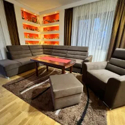 Brązowy, tapicerowany narożnik z poduchami, stolik kawowy z czerwoną serwetą i fotel z pufą na brązowym dywanie we wnętrzu z półkami wypełnionymi dekoracjami.