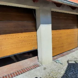 Dwa garaże z segmentowymi bramami w kolorze drewna, jedna brama lekko uchylona, widoczna kostka brukowa i fragment ogrodu w tle.