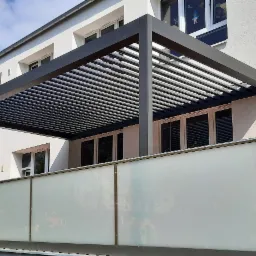 Nowoczesna pergola z poziomymi lamelami w kolorze antracytowym, zamontowana nad balkonem z matowym szkłem, widok z zewnątrz na tle jasnej elewacji budynku i zieleni drzew