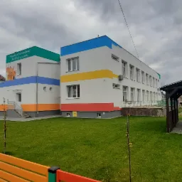 Nowoczesny budynek przedszkola z kolorowymi akcentami, widoczne okna, ogrodzenie z desek w barwach tęczy i drewniana altana z ławkami na trawniku.